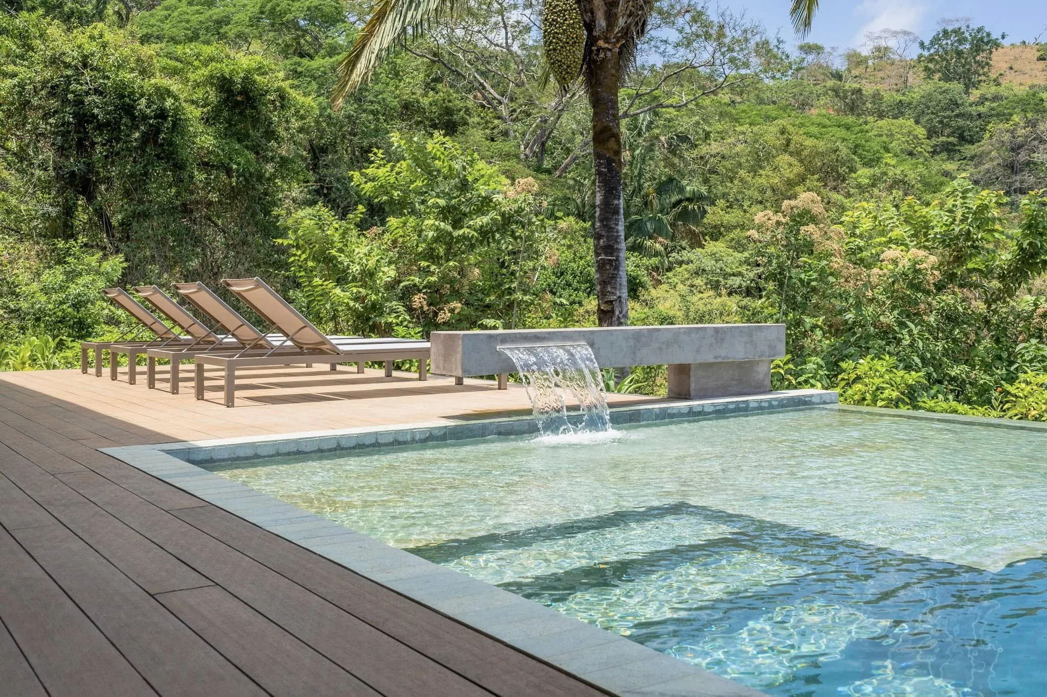 An outdoor swimming pool with clear water, surrounded by a wooden deck with four lounge chairs, set against a lush green tropical forest and blue sky.