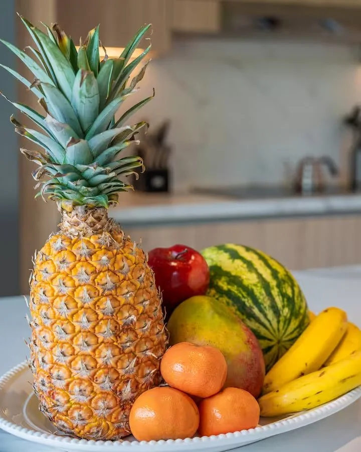 Assorted fresh fruits including a pineapple, bananas, apples, oranges, a melon, and a pear on a white tray.