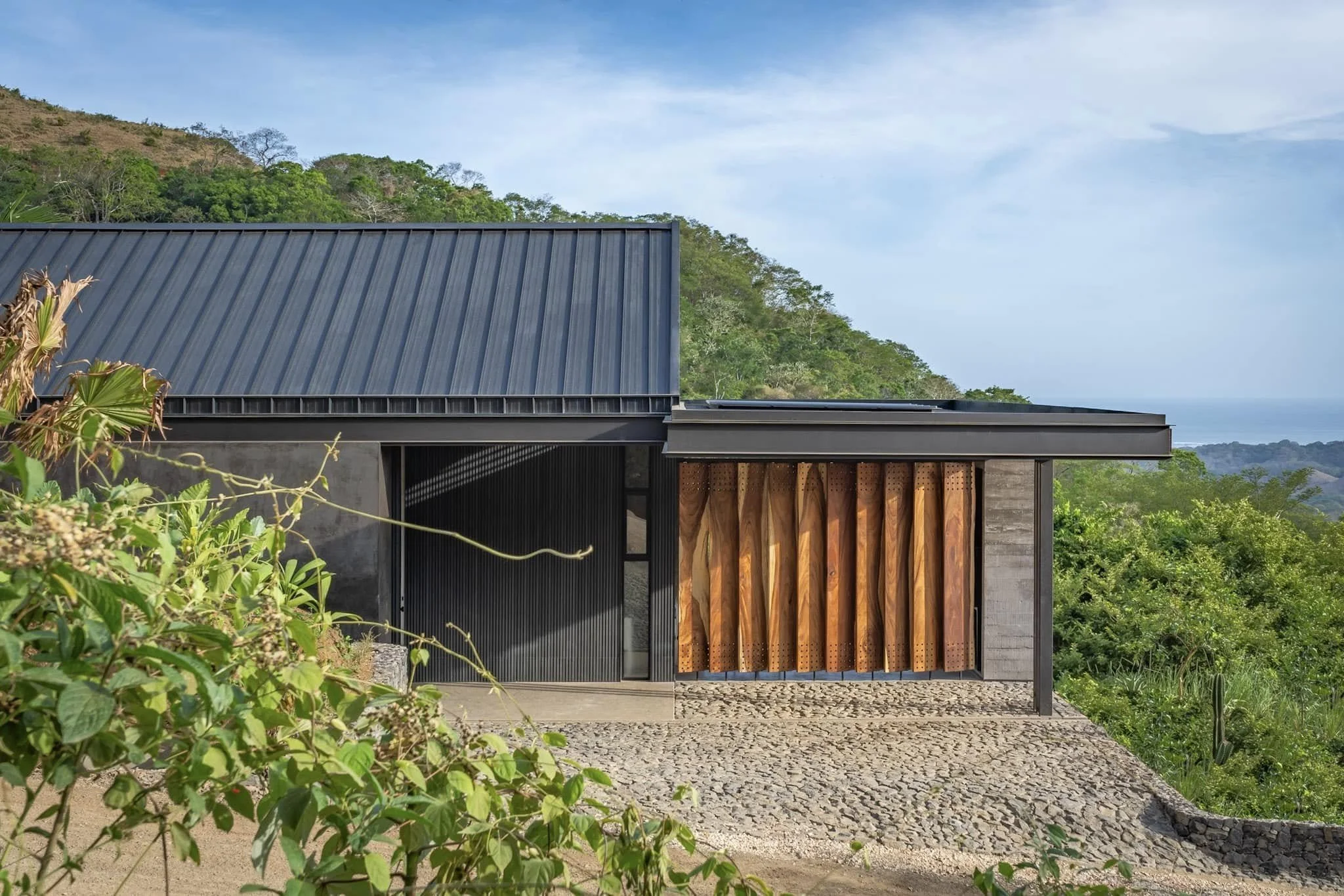 Modern house with a metal roof, wooden panel accents, and large sliding glass doors, situated on a hillside with lush green trees and hills in the background.