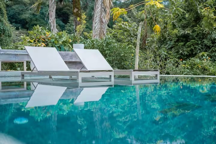 Empty pool with two lounge chairs and lush green tropical vegetation in the background.
