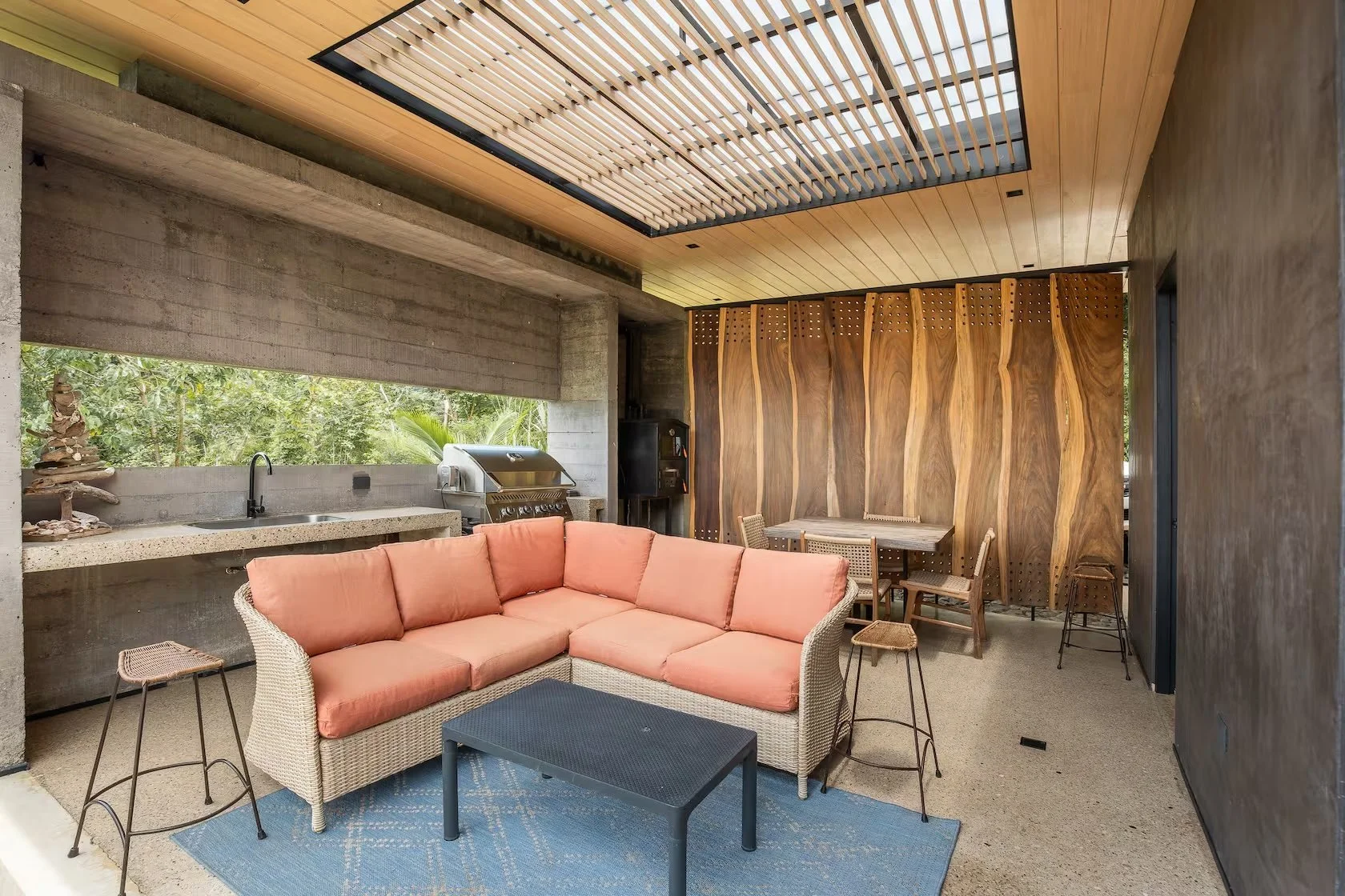 Indoor living space with pink cushioned outdoor sofa, black coffee table, and stools, open kitchen area with concrete countertop, sink, and grill, wood and concrete walls, and a wooden ceiling with a skylight.