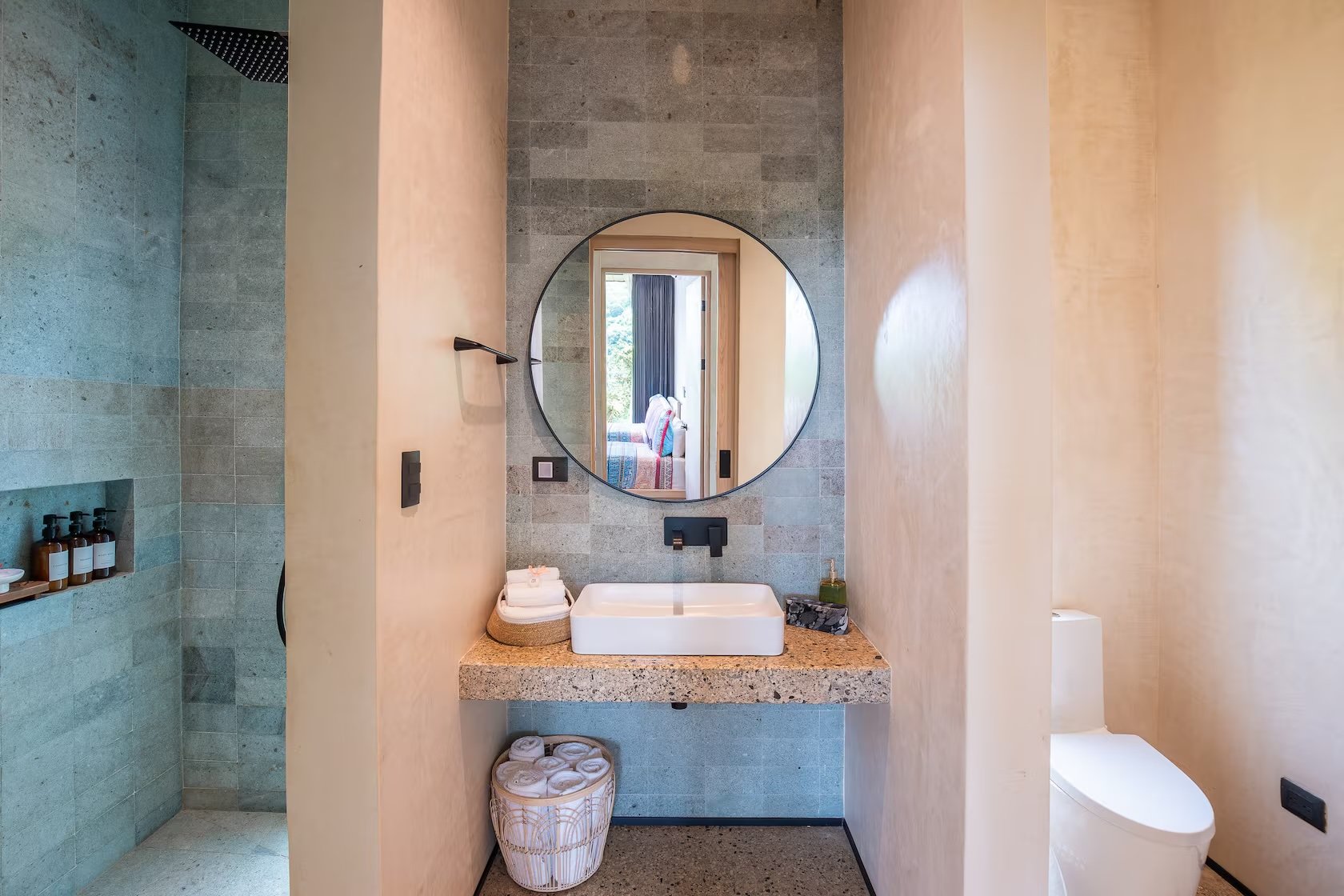 A modern bathroom with a circular mirror above a rectangular sink, a view into a bedroom through a doorway, and a shower on the left with toiletries on shelves.