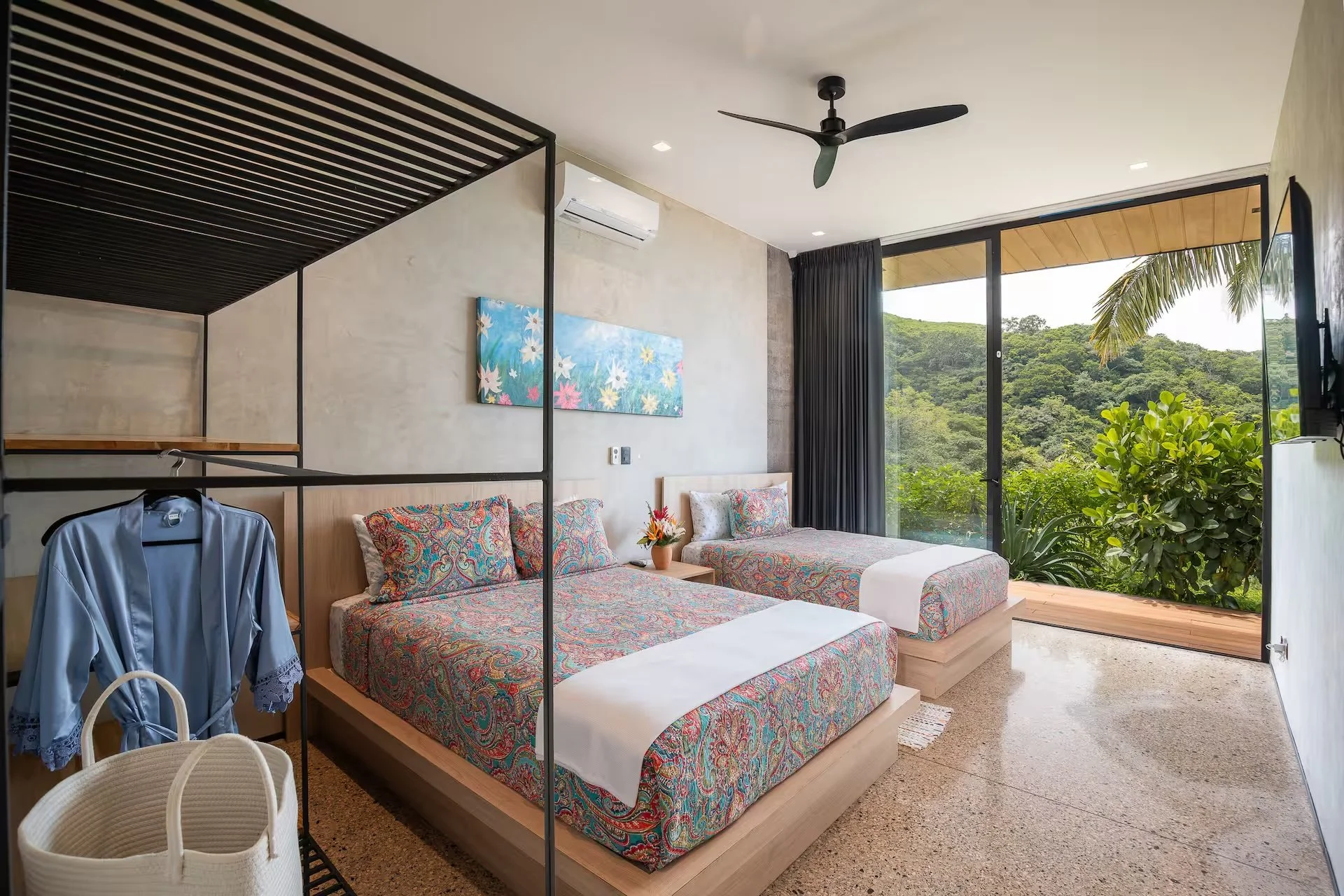 Two beds with colorful paisley bedspreads in a modern room with large glass doors showing outdoor greenery and a palm tree, a ceiling fan, an air conditioning unit, a decorative painting on the wall, and a small wooden table with flowers.