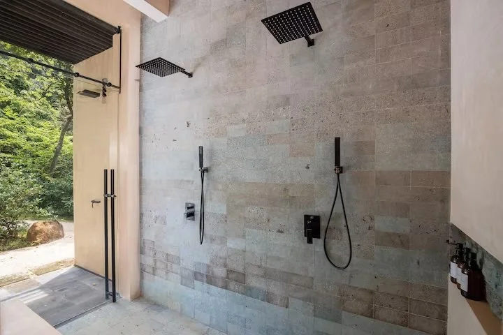Outdoor shower area with two black rainfall showerheads mounted on a tiled wall, black handheld shower fixtures, and a doorway leading to an outdoor space with trees and rocks.