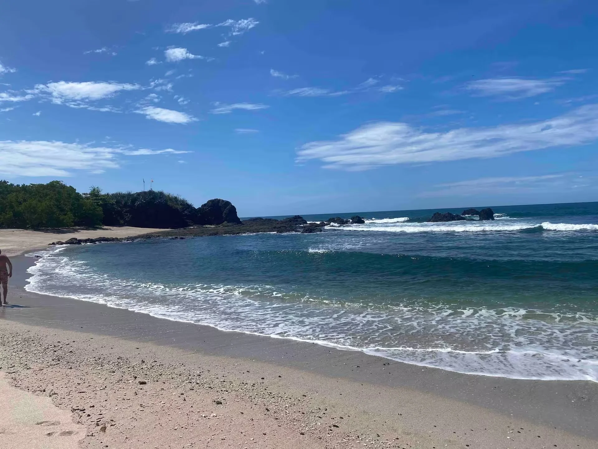 A sandy beach with footprints leading to the water, waves crashing on rocks, green shrubbery, and palm trees on a small hill with flags, under a bright blue sky with scattered clouds.