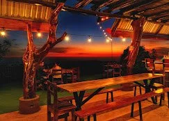 Outdoor dining area at sunset with wooden tables and benches under a canopy, overlooking a scenic landscape.