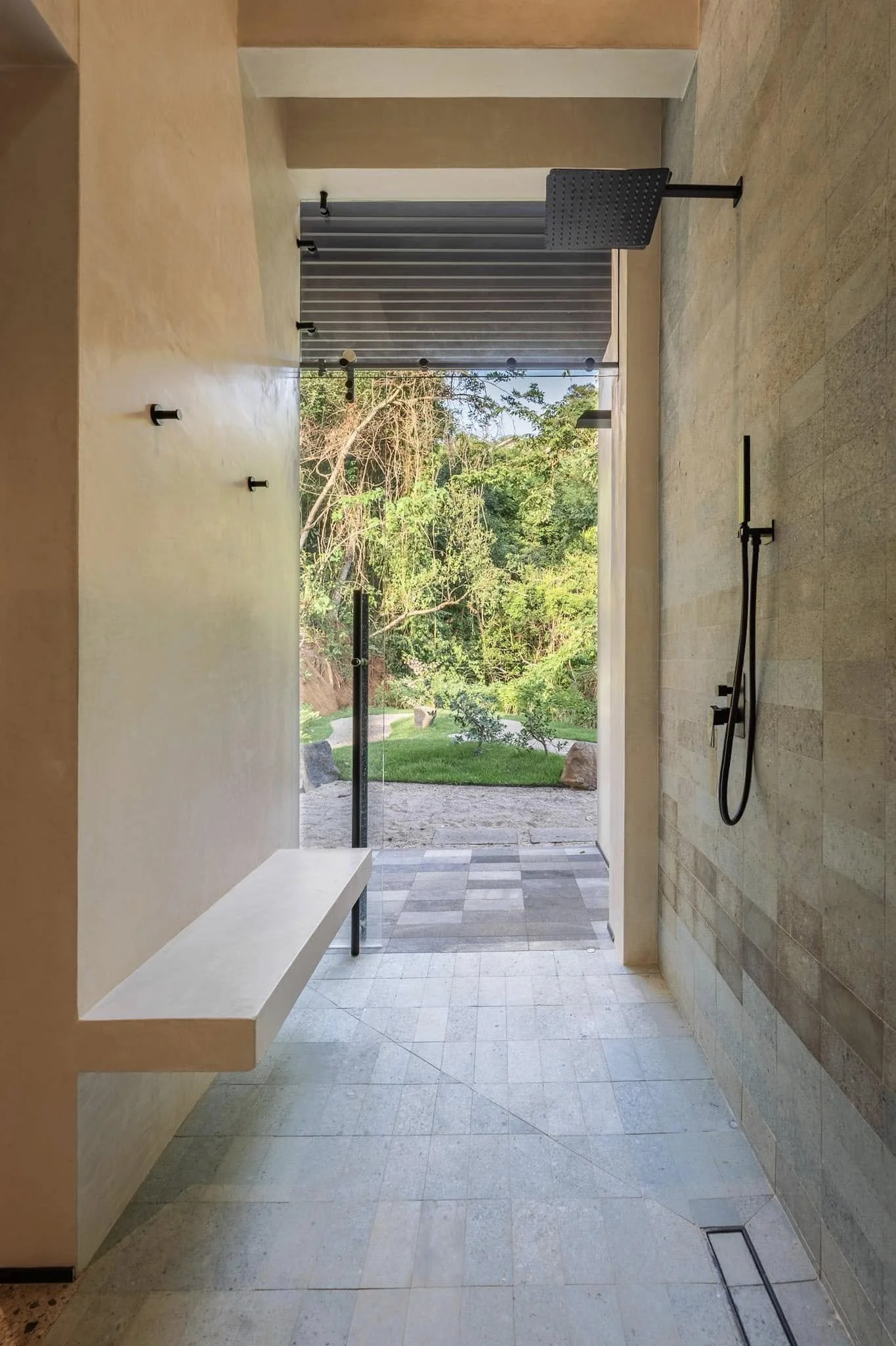 Modern interior space with beige tiled floor and walls, featuring a large open doorway leading to an outdoor area with greenery and trees. There are some black fixtures and a black chair inside the room, with a long angled white bench along one wall and a floor vent near the bottom right corner.