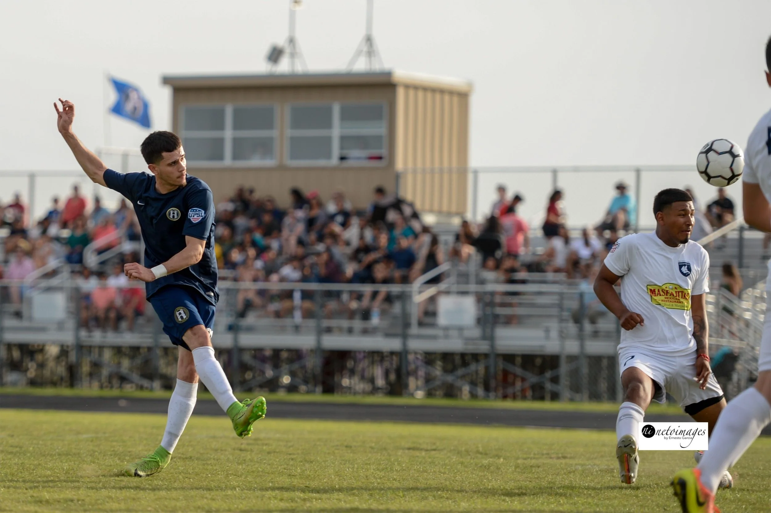 Waco SC Soccer players on the field during a match, with one player in a navy jersey and green cleats kicking the ball and another player in a white jersey and black cleats nearby, spectators in the background.
