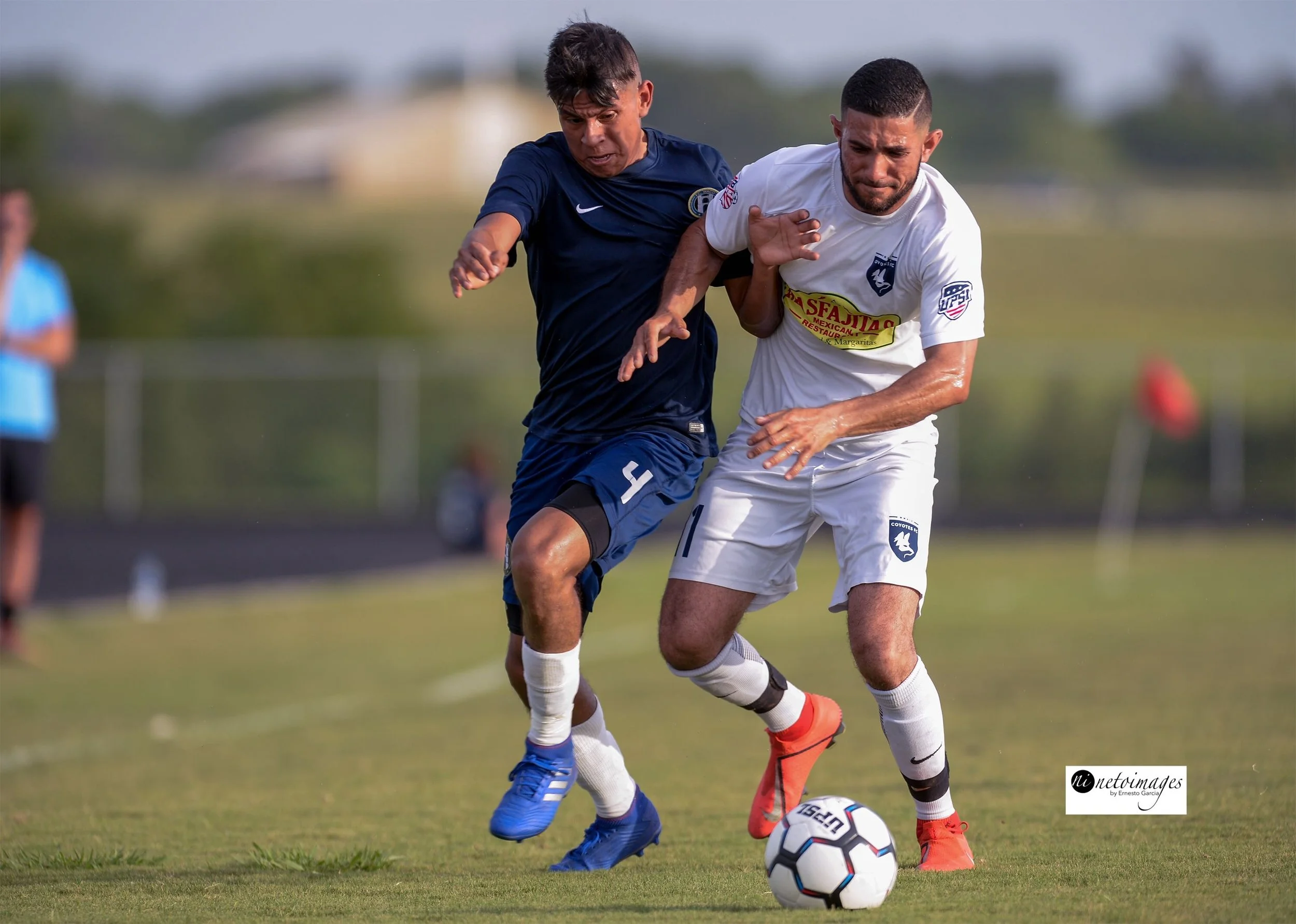Two male soccer players competing for the ball on the field during a game, one in a dark blue Waco Soccer Club jersey and the other in a white jersey.