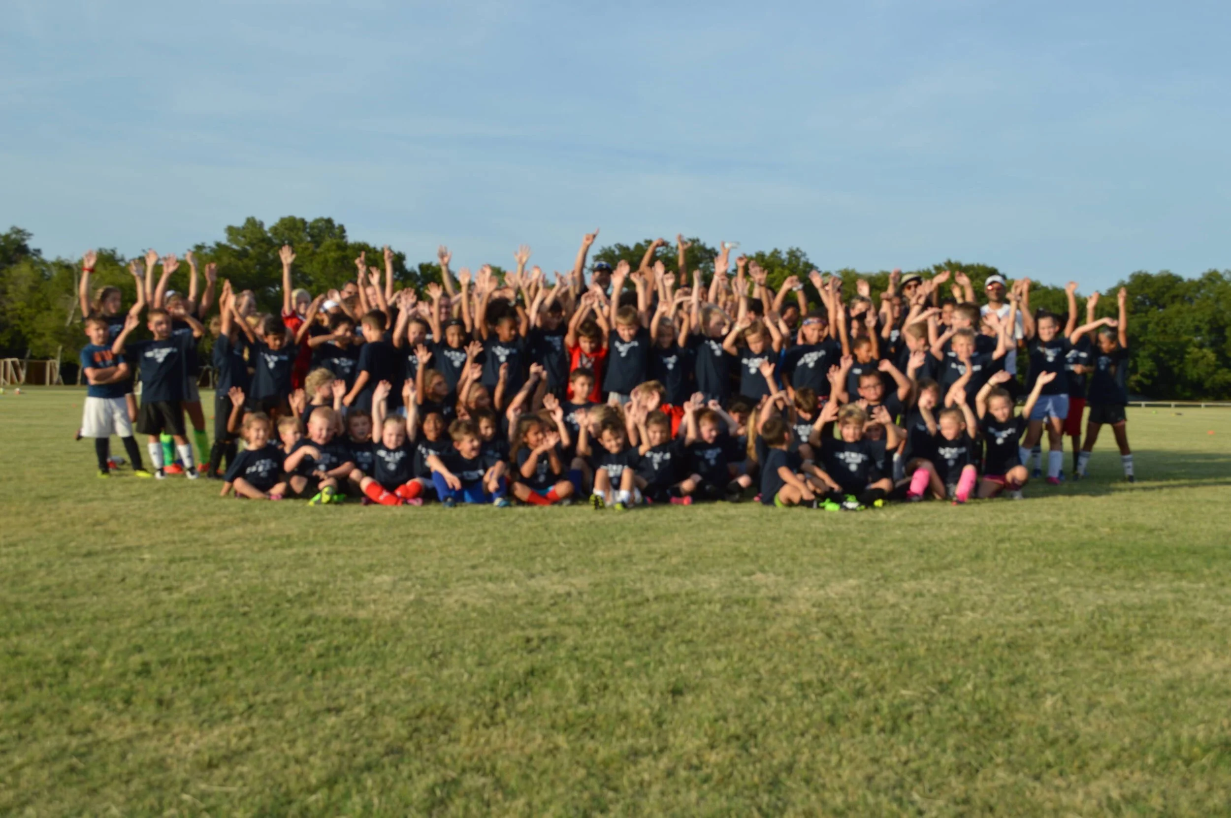 A large group of children on a sports field, participating in a Waco Soccer Club camp. Many children are wearing matching navy blue waco sc shirts, some are seated in front, while others are standing and raising their hands or making gestures.