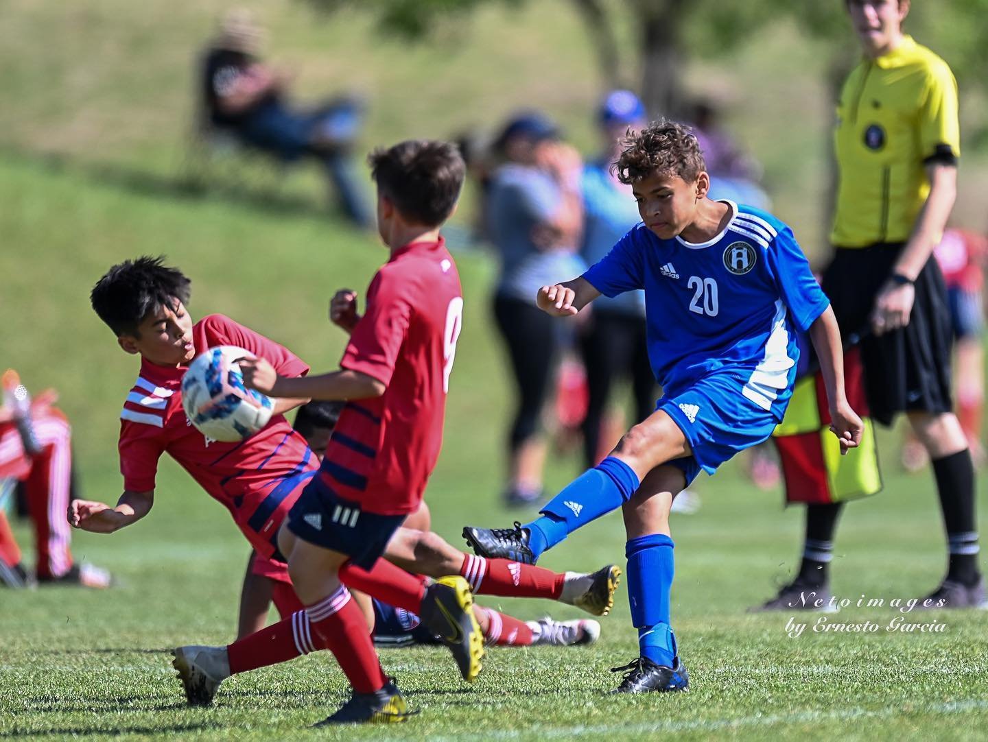 Young boys playing soccer on a grassy field, with one boy in a Waco SC Uniform kicking a soccer ball as two boys in red uniforms attempt to block him. 