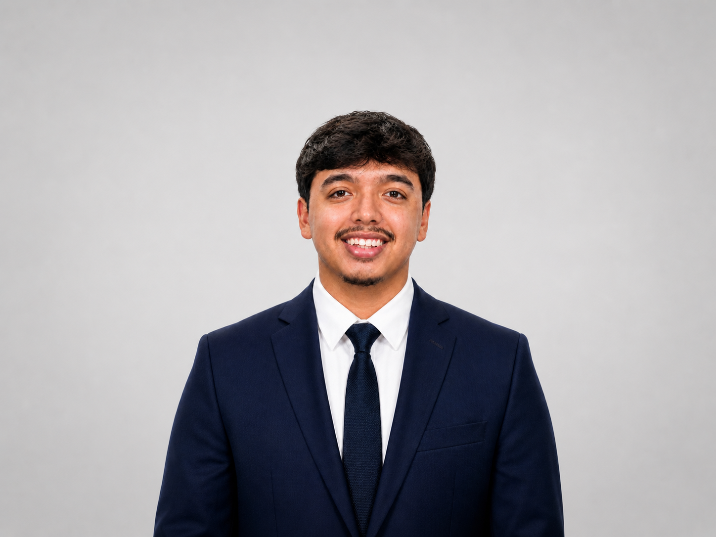 A young man in a navy blue suit, white shirt, and navy tie, smiling at the camera against a plain light gray background. Waco SC business development intern.