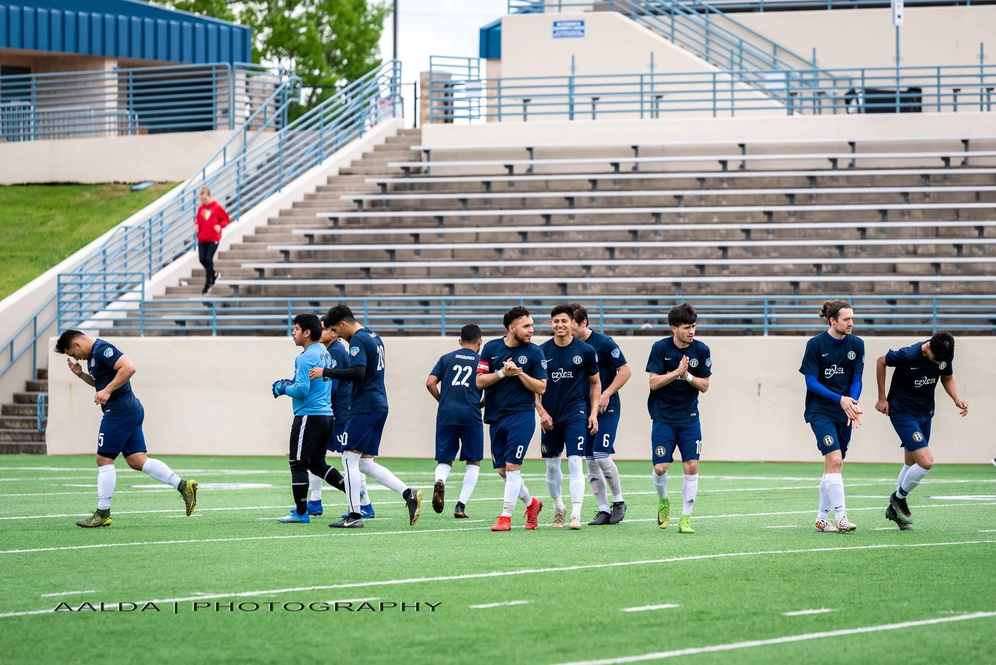 A group of Waco SC soccer players in blue uniforms walking on a soccer field with a stadium in the background.
