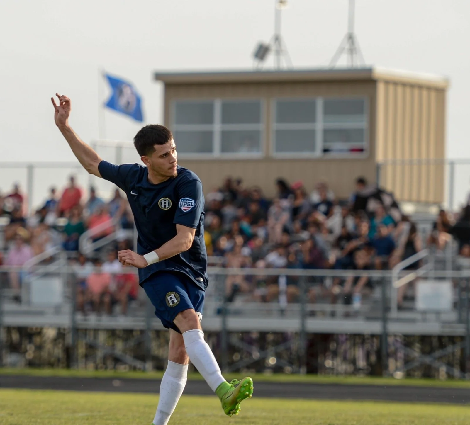 A waco soccer club player in navy uniform with a badge and a patch on the sleeve, kicking a ball on a field with waco sc fans in the background.