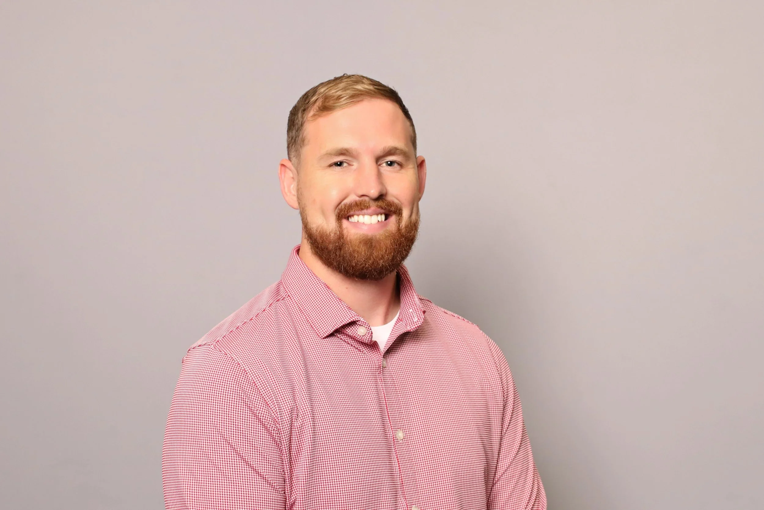 A man with a reddish beard and short hair, wearing a pink checkered shirt, smiling, standing against a plain light gray background. Government Relations for Waco Soccer Club.