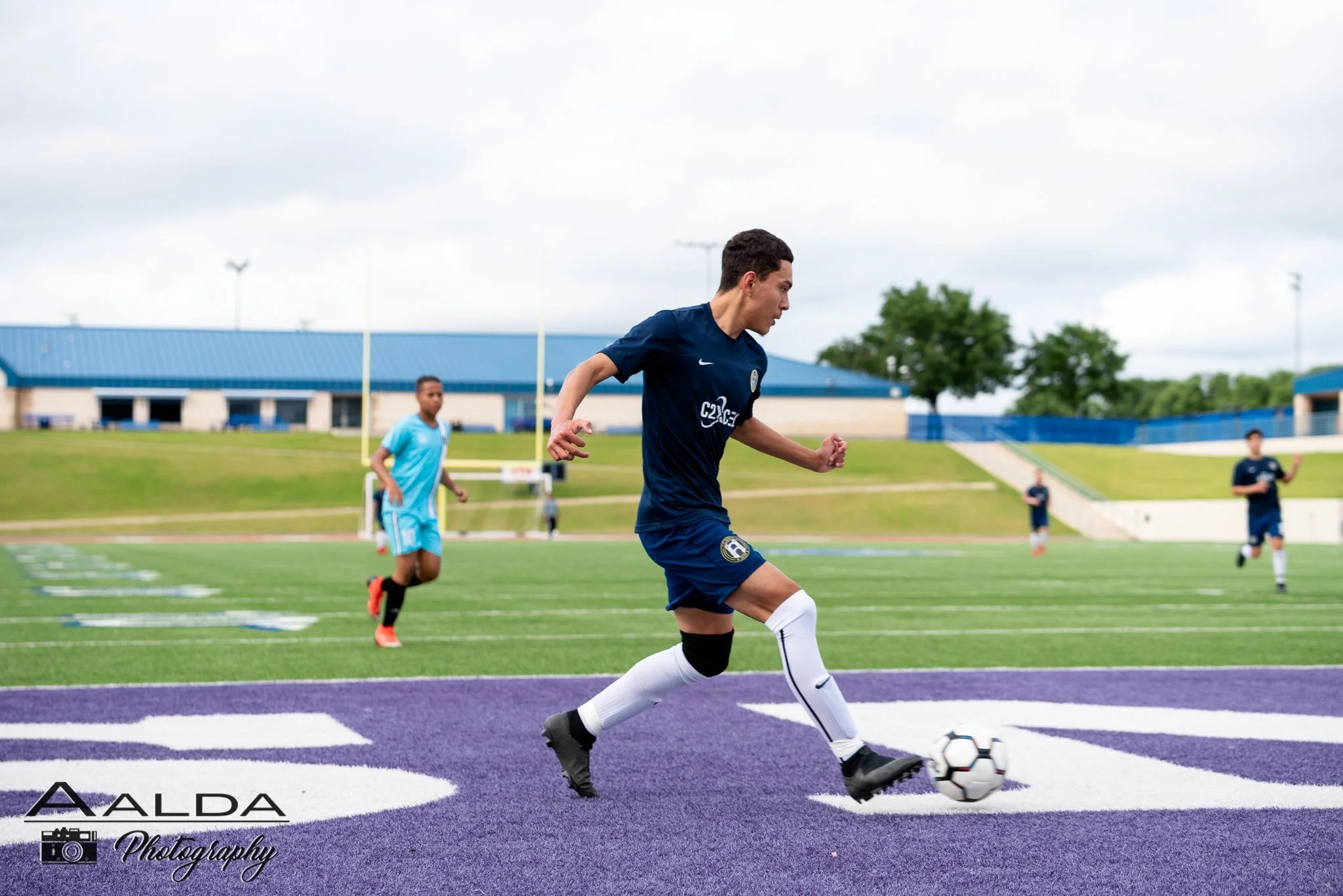 A young man in a blue Waco SC soccer uniform kicking a soccer ball on a colorful outdoor field during daytime. Other players and a building are visible in the background.