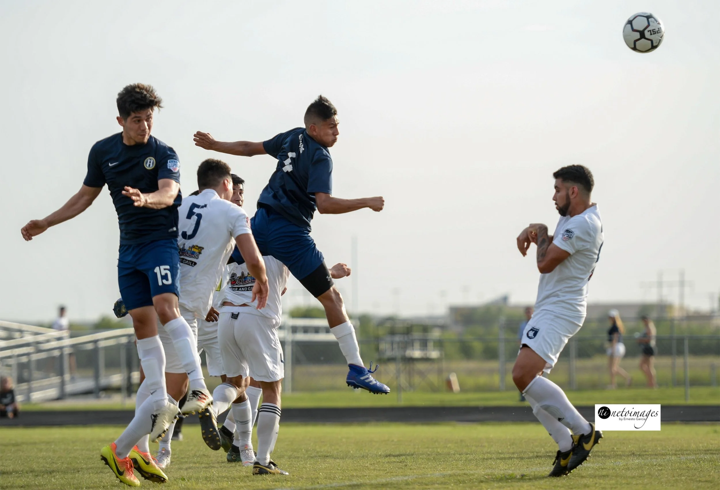 Waco SC Soccer players in blue and white uniforms jumping and heading a soccer ball during a game on a grassy field.