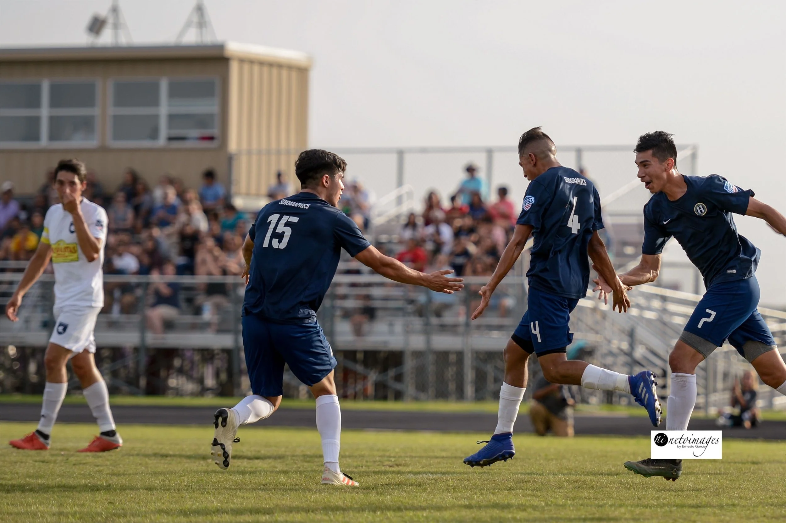 Waco Soccer Club players celebrating on the field during a match, with spectators in the stands in the background.