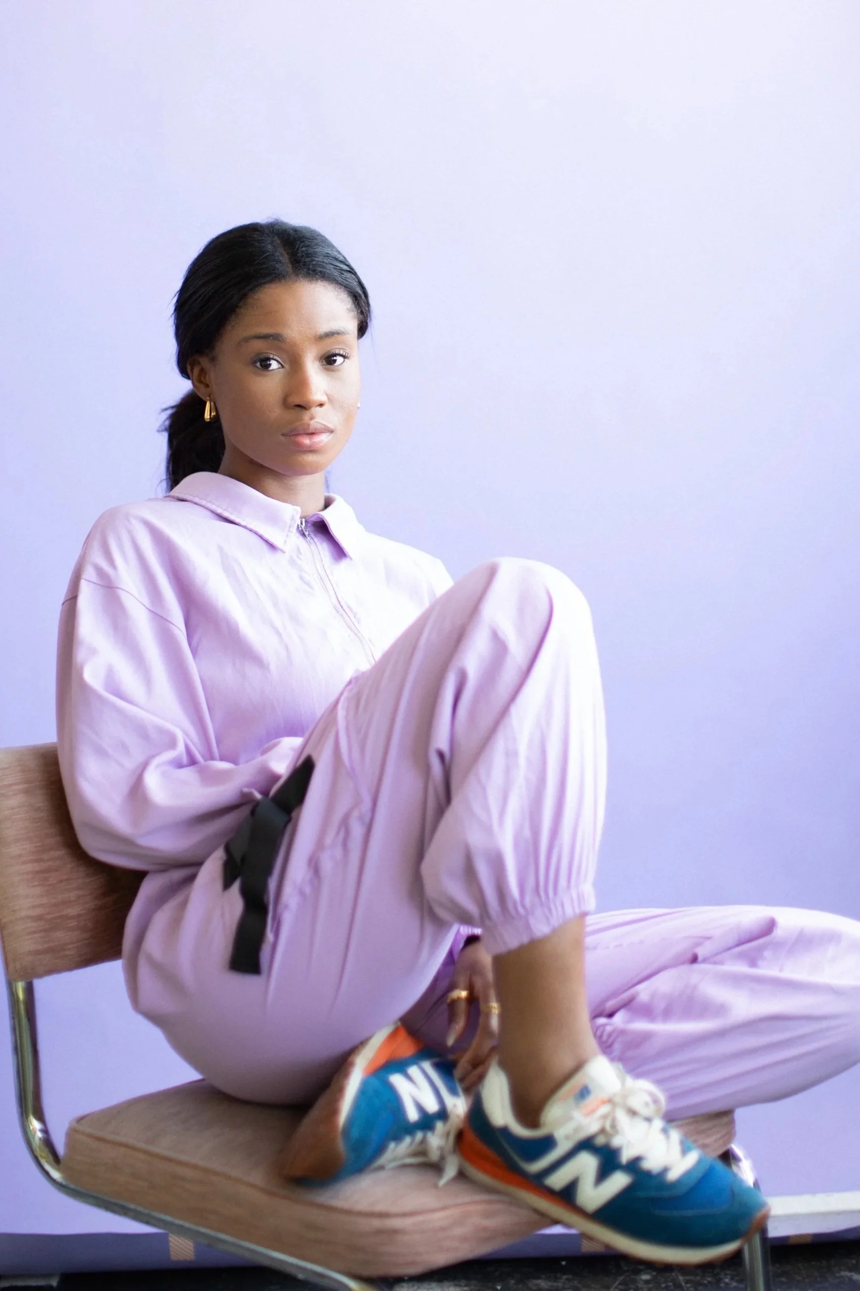 This image is of Adeyinka Akinrinade the founder of veritas productions. She is sat down, wearing a purple jumpsuit with a light purple background.
