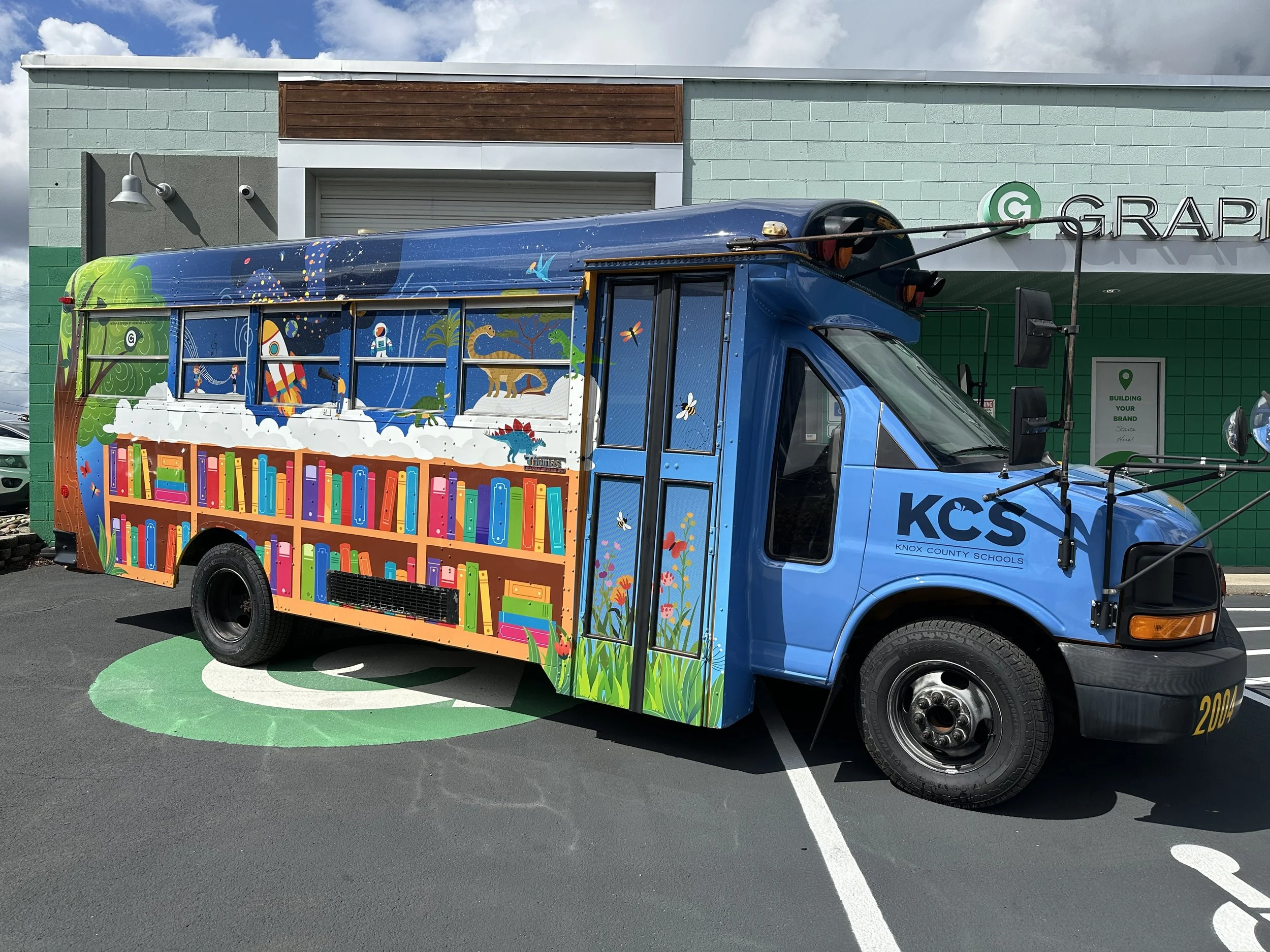 A colorful school bus decorated with books, dinosaurs, and space-themed illustrations parked in a parking lot in front of a building with a sign that says 'GRA' and a green circle logo.