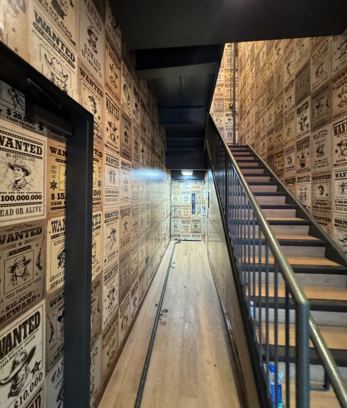 An indoor staircase area with wallpapered walls featuring vintage wanted posters from the Old West, and a narrow wooden floor with metal railings on the stairs.