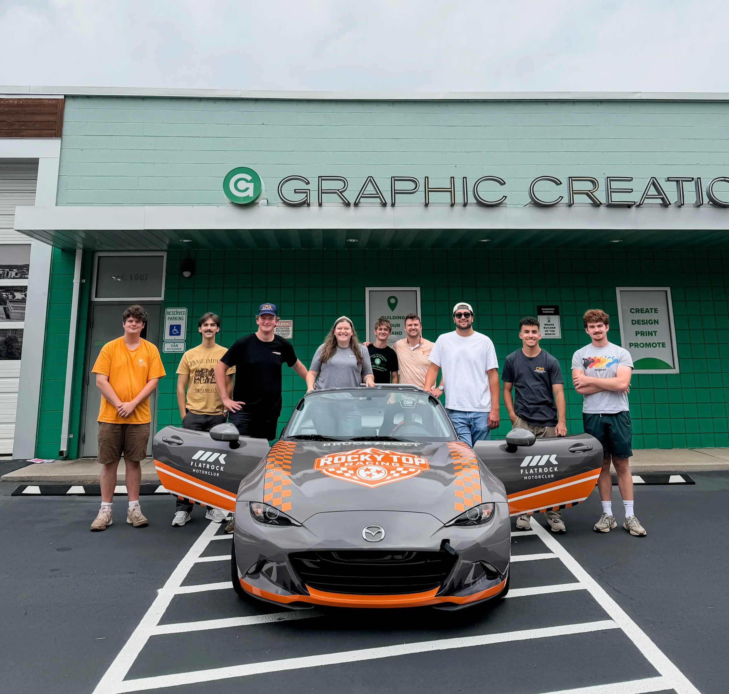 A group of ten people standing in front of a green building with the sign 'GRAPHIC CREATION' and a small race car with a black and orange design in a parking lot.
