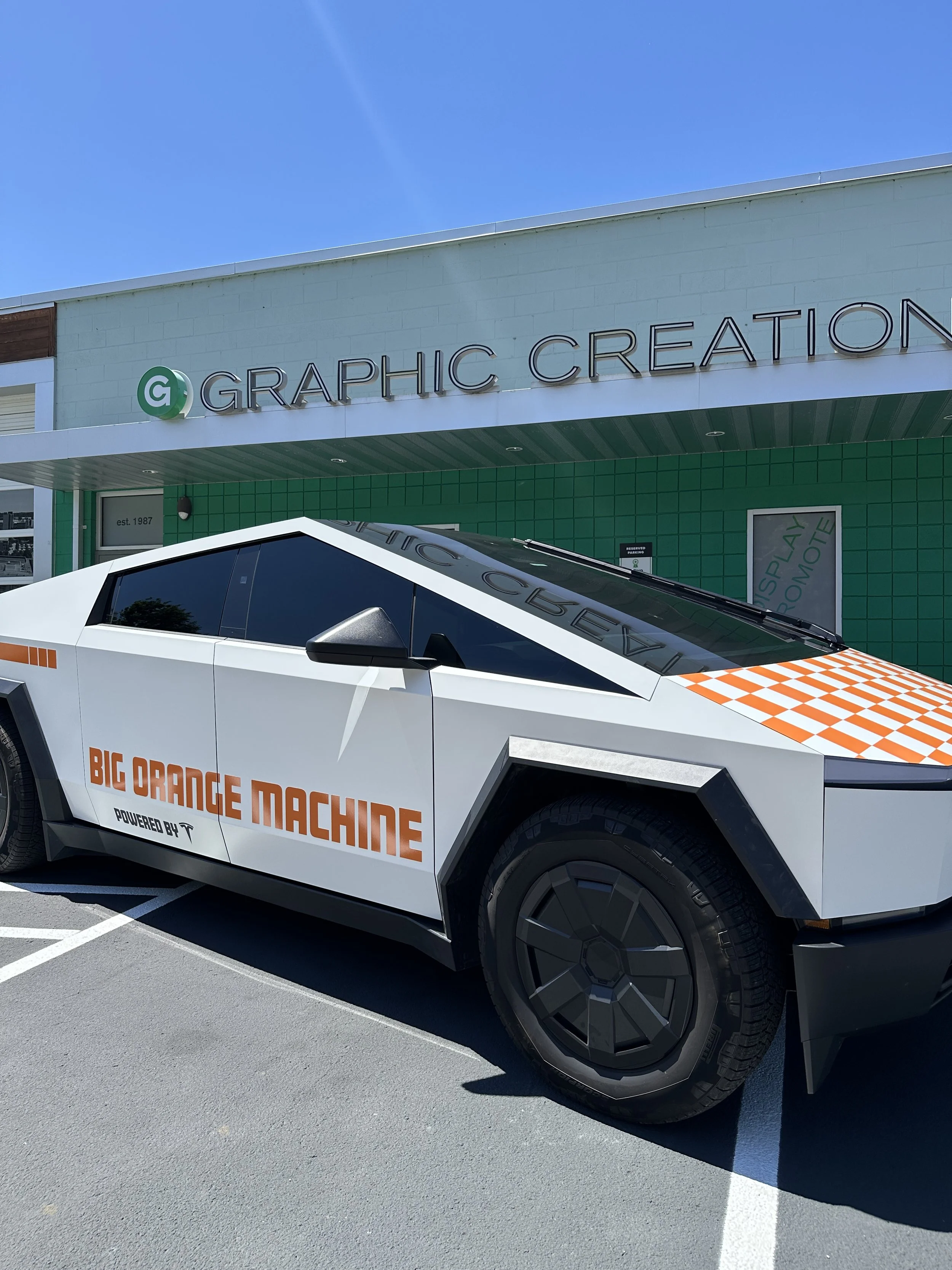 A futuristic white car with black wheels parked in a parking lot with a sign for Graphic Creation and Big Orange Machine in the background.