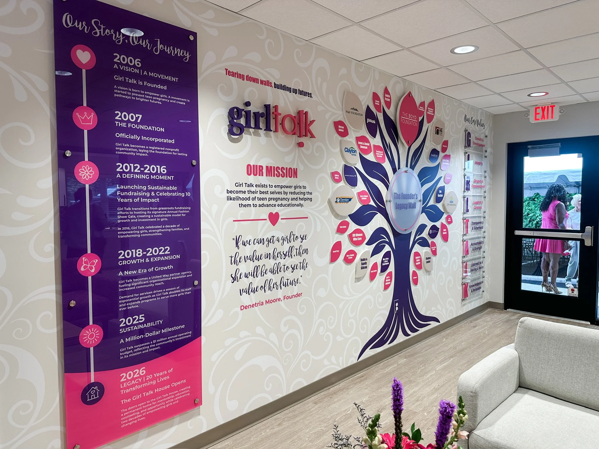 Interior wall display highlighting Girl Talk organization history, mission, and milestones from 2006 to 2026, with a decorative tree graphic, flowers in the foreground, and an exit door on the right.
