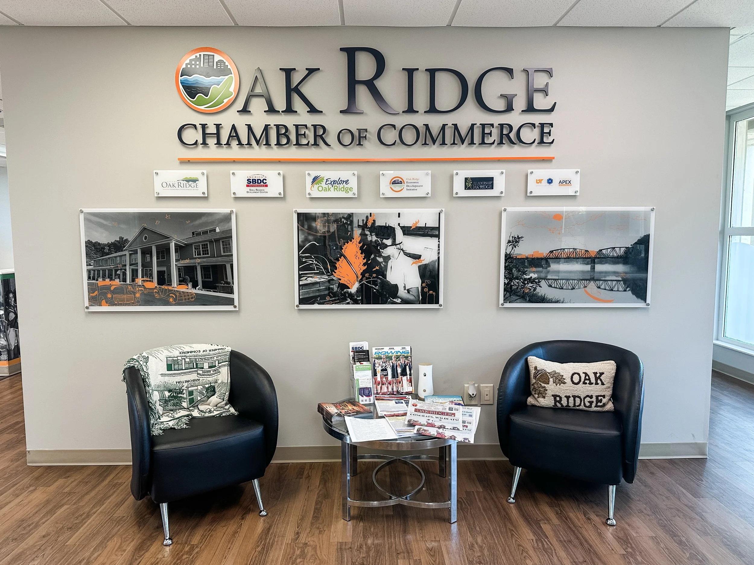 The image shows a reception or waiting area for the Oak Ridge Chamber of Commerce. There are two black chairs with cushions, a small round table with magazines, pamphlets, and flyers, and a decorative pillow. The wall behind displays a large sign wit