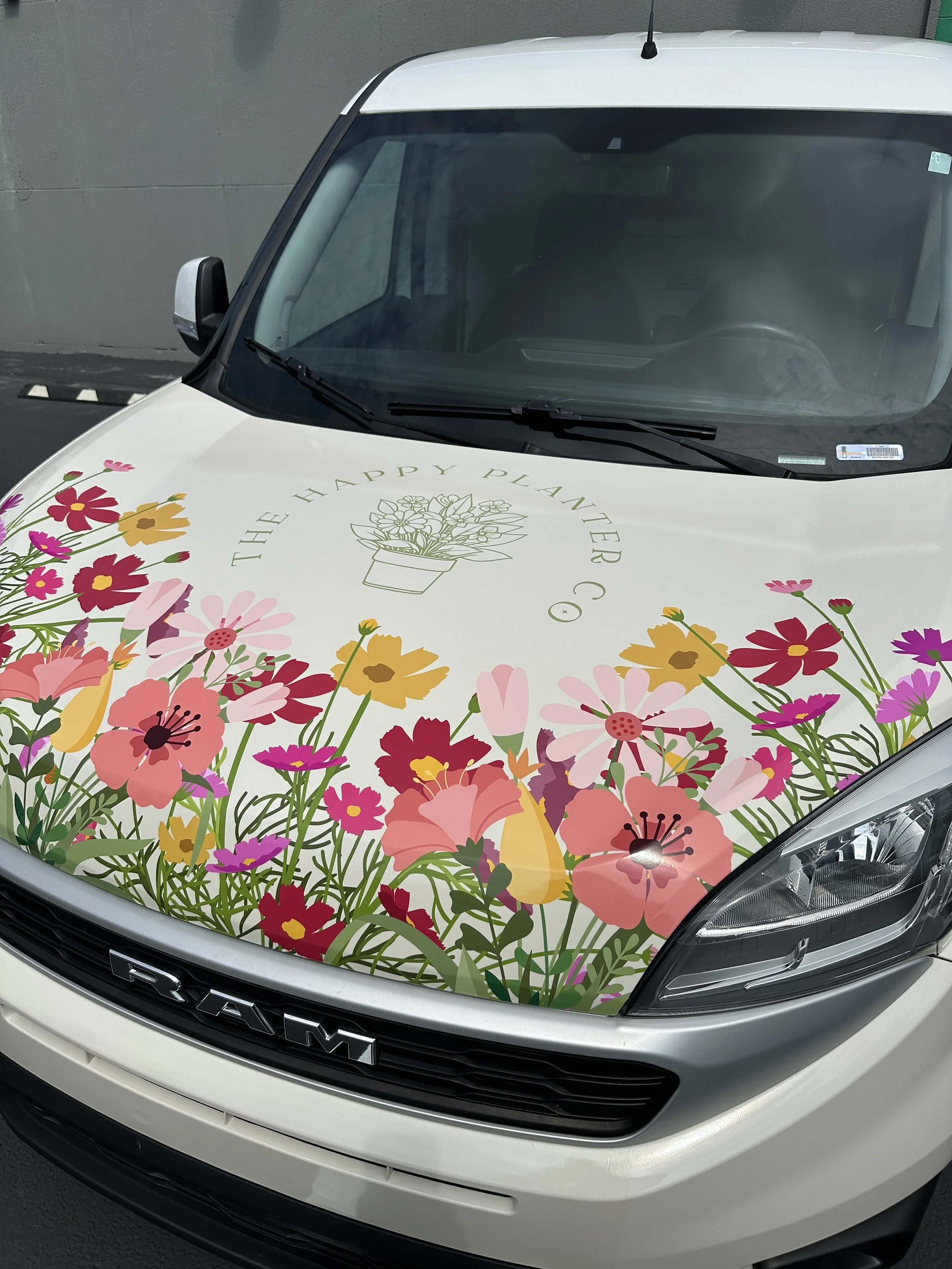A white Ram pickup truck with a floral design on the hood, featuring various pink, yellow, and purple flowers and the phrase 'The Happy Planter Co.' with an illustration of a flower pot.