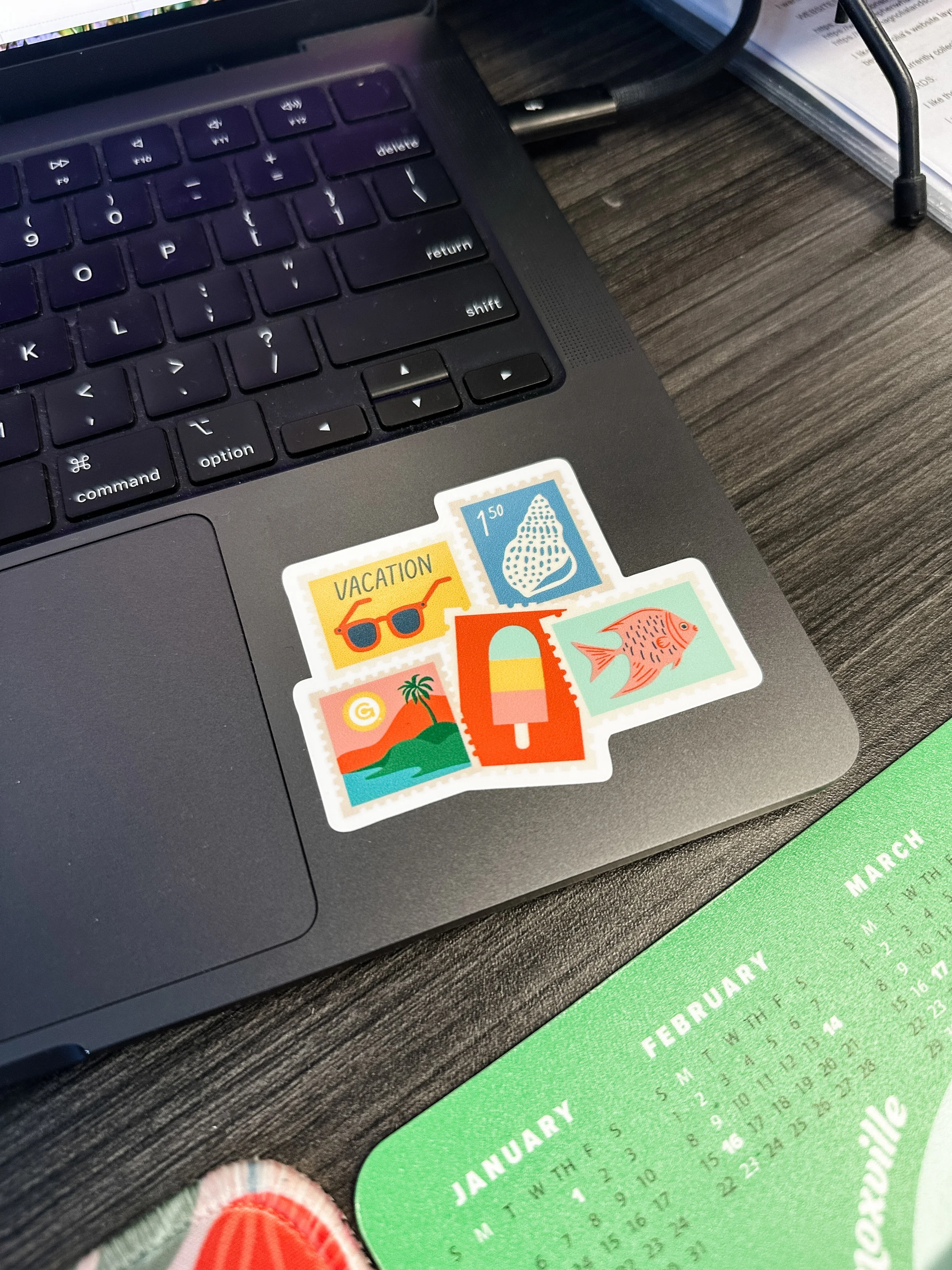 Close-up of a laptop keyboard on a wooden table, with colorful travel-themed stickers on the lower right corner of the laptop. A green calendar displaying the month of January is also partially visible at the bottom of the image, and a person's foot 