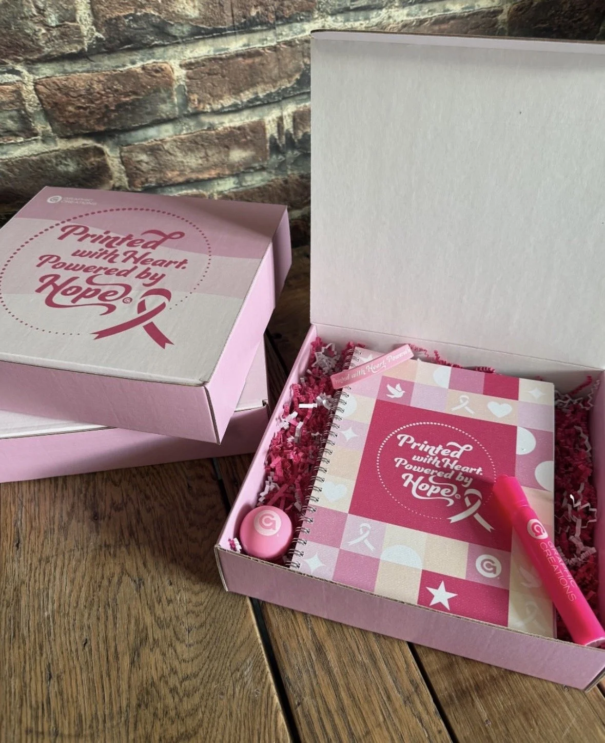 Open pink box with breast cancer awareness theme, containing a pink notebook titled "Printed with Heart: Powered by Hope," a pink marker, a pink round object, and red shredded paper on a wooden surface with a brick wall background.