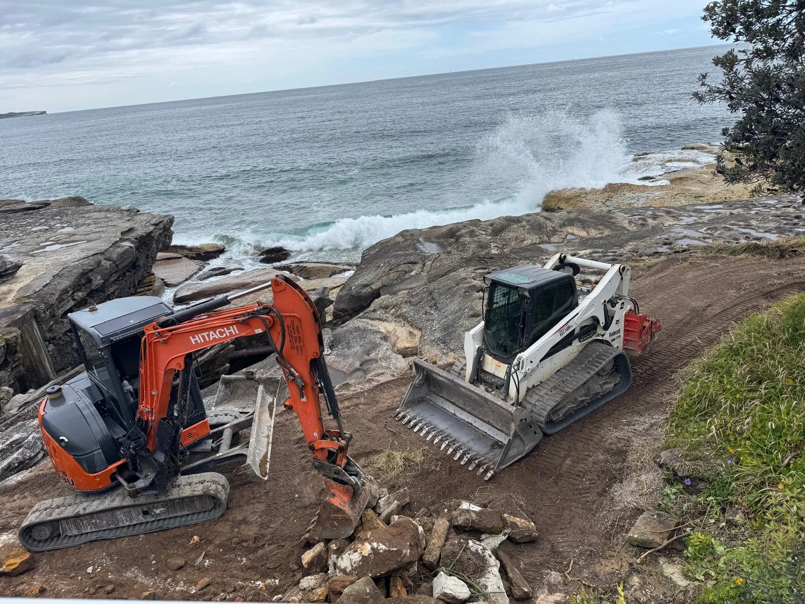 Two construction mini excavators on a dirt path near a rocky coastline in Australia
