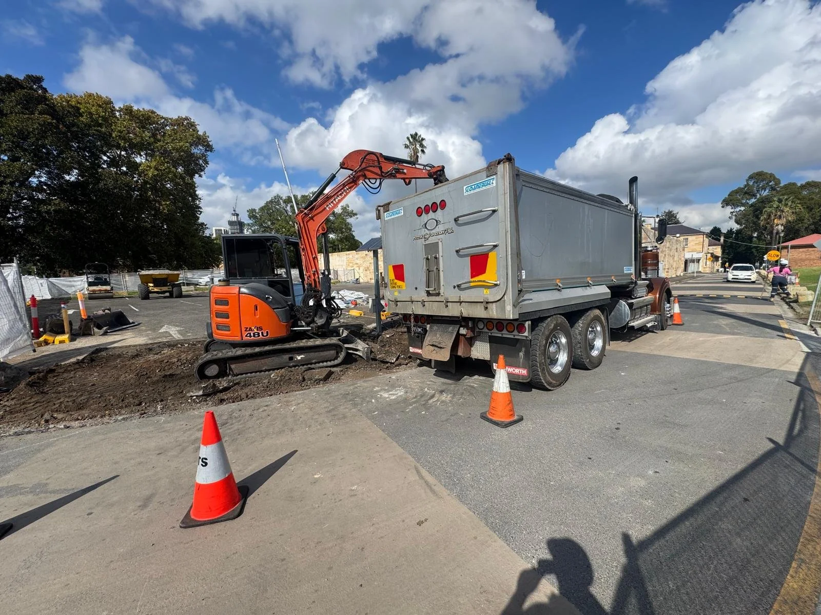 A construction site on a street with an orange excavator and a grey dump truck. 