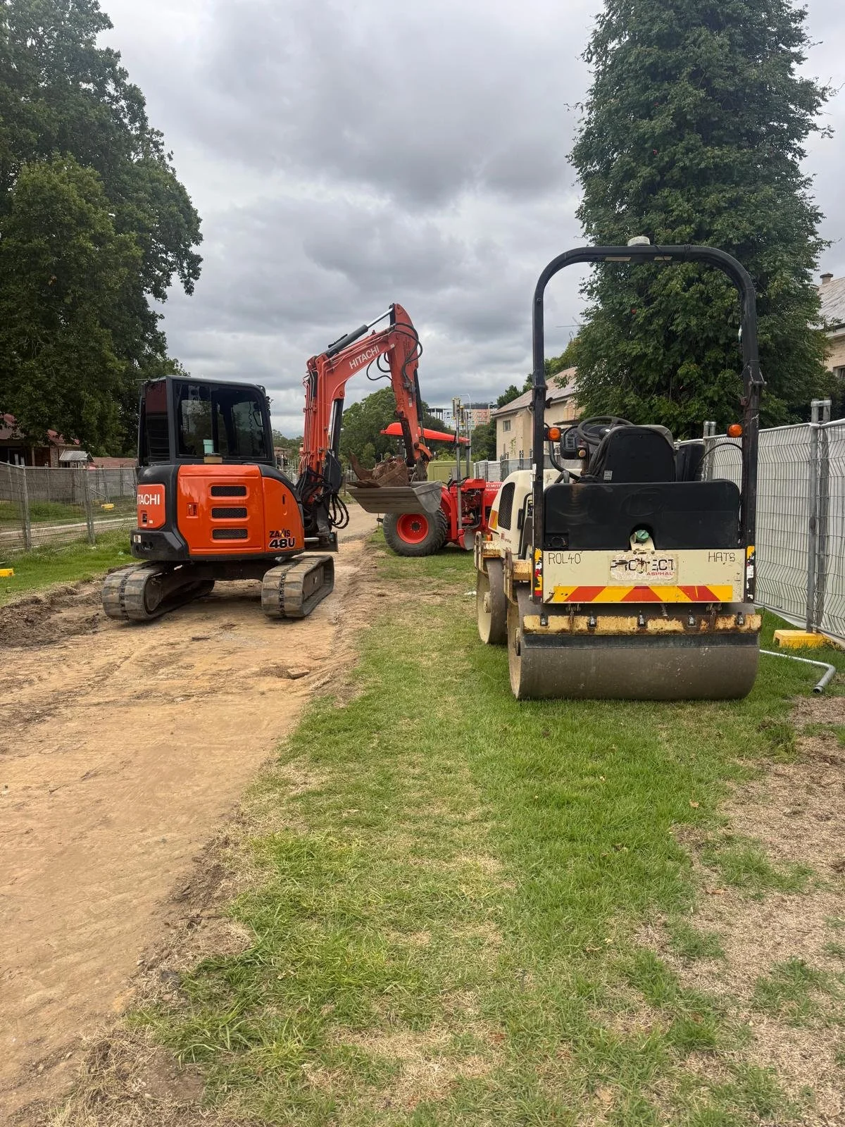 Construction site with three different types of equipment: a small orange excavator, a red loader with a bucket, and a black and yellow road roller