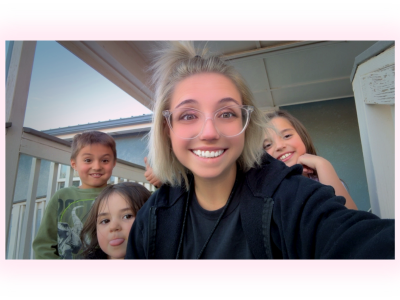 A young woman with glasses and blonde hair taking a close-up selfie with four children, all smiling and posing for the camera on a porch.