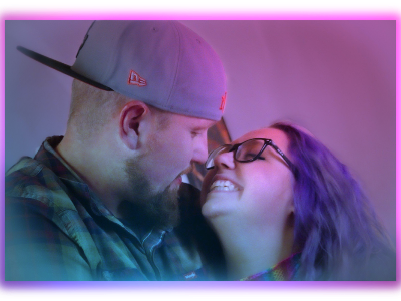 A man and woman are close to each other, smiling and touching foreheads, with a pinkish-purple background and lighting.