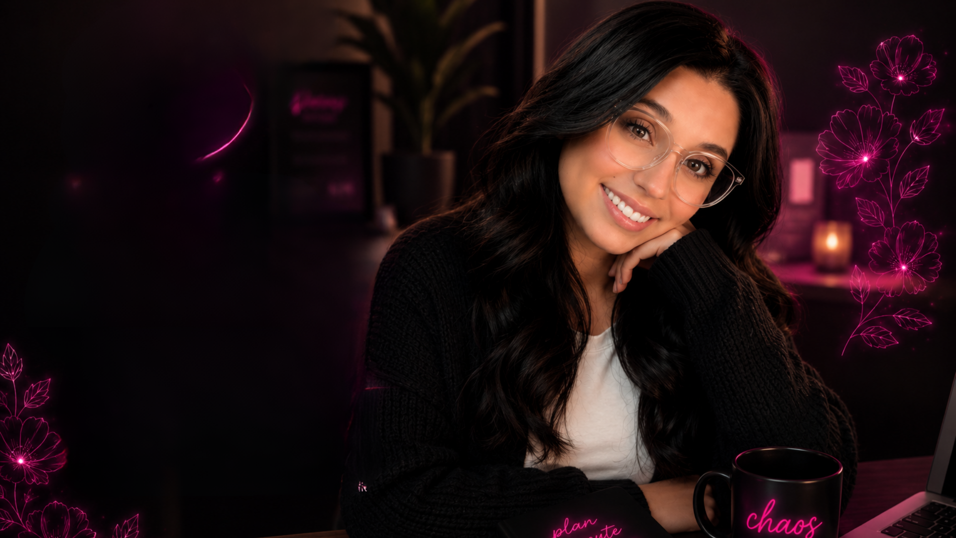 A young woman with long dark hair, glasses, and a black sweater, smiling and resting her chin on her hand in a dimly lit room with pink neon floral decorations.