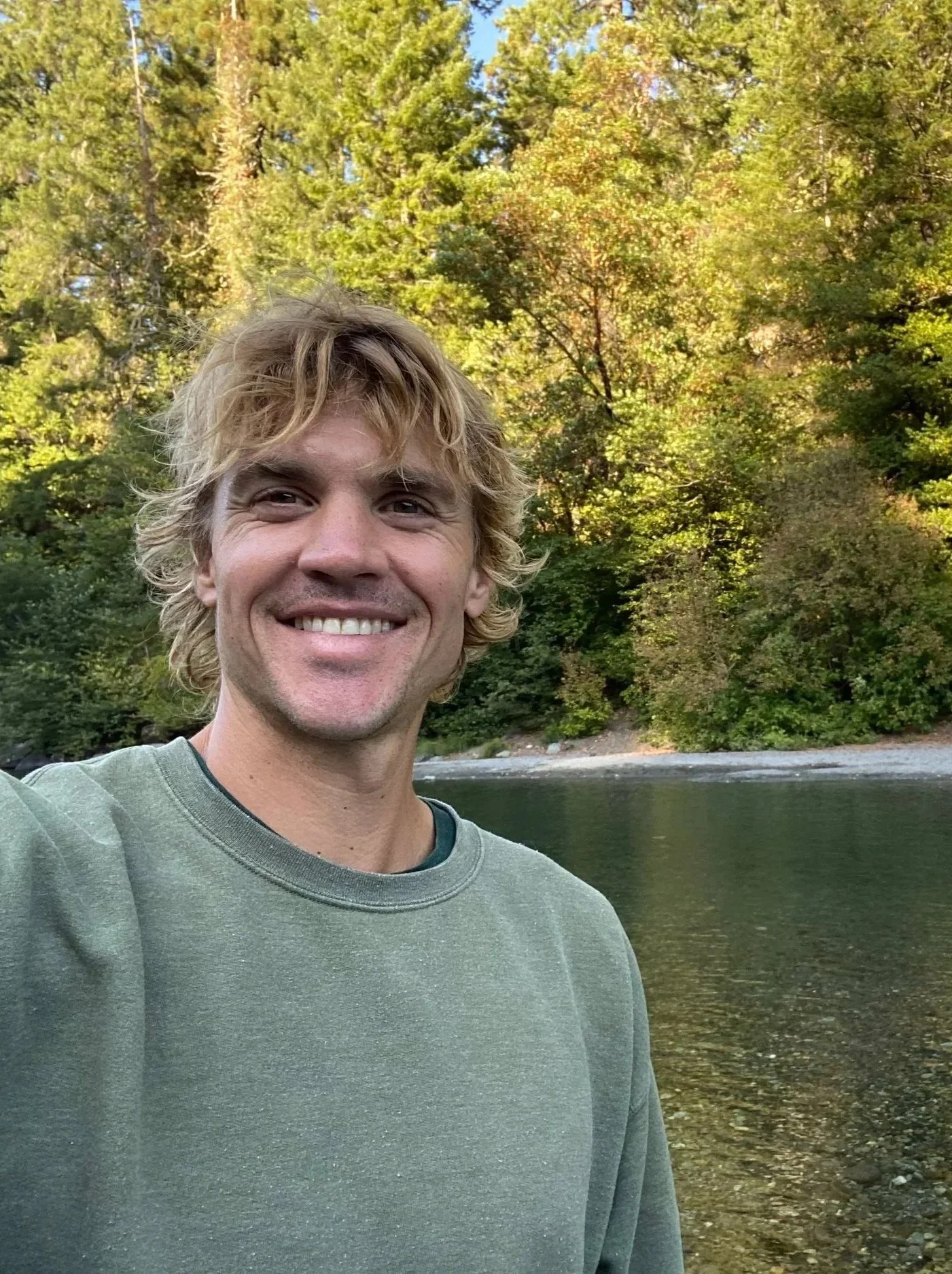 A smiling man, Nathan Toben, with blonde hair wearing a green sweatshirt outdoors by a body of water, with trees and autumn foliage in the background.