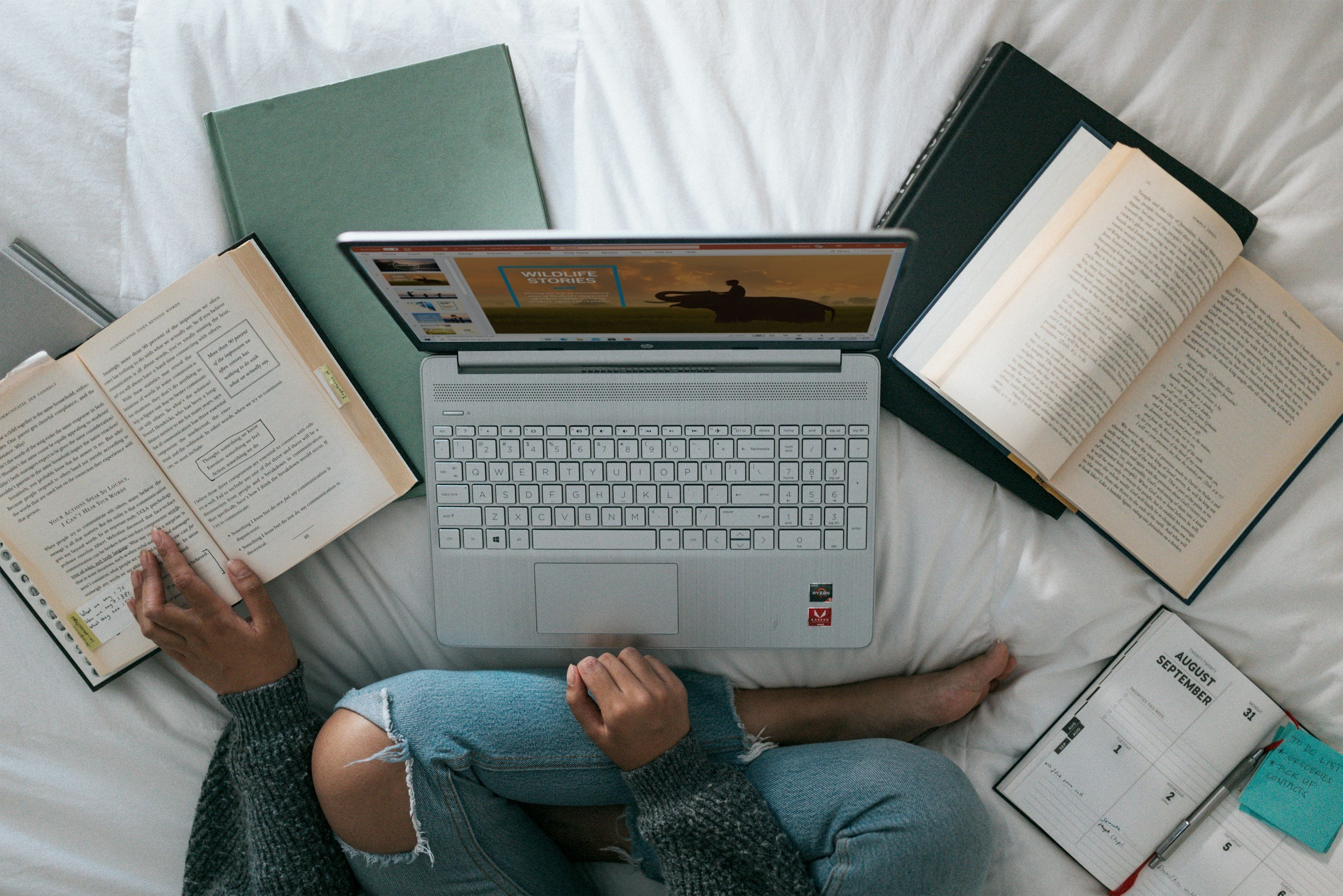 Person sitting on a bed reading a book with an open laptop in front of them, surrounded by notebooks and papers.