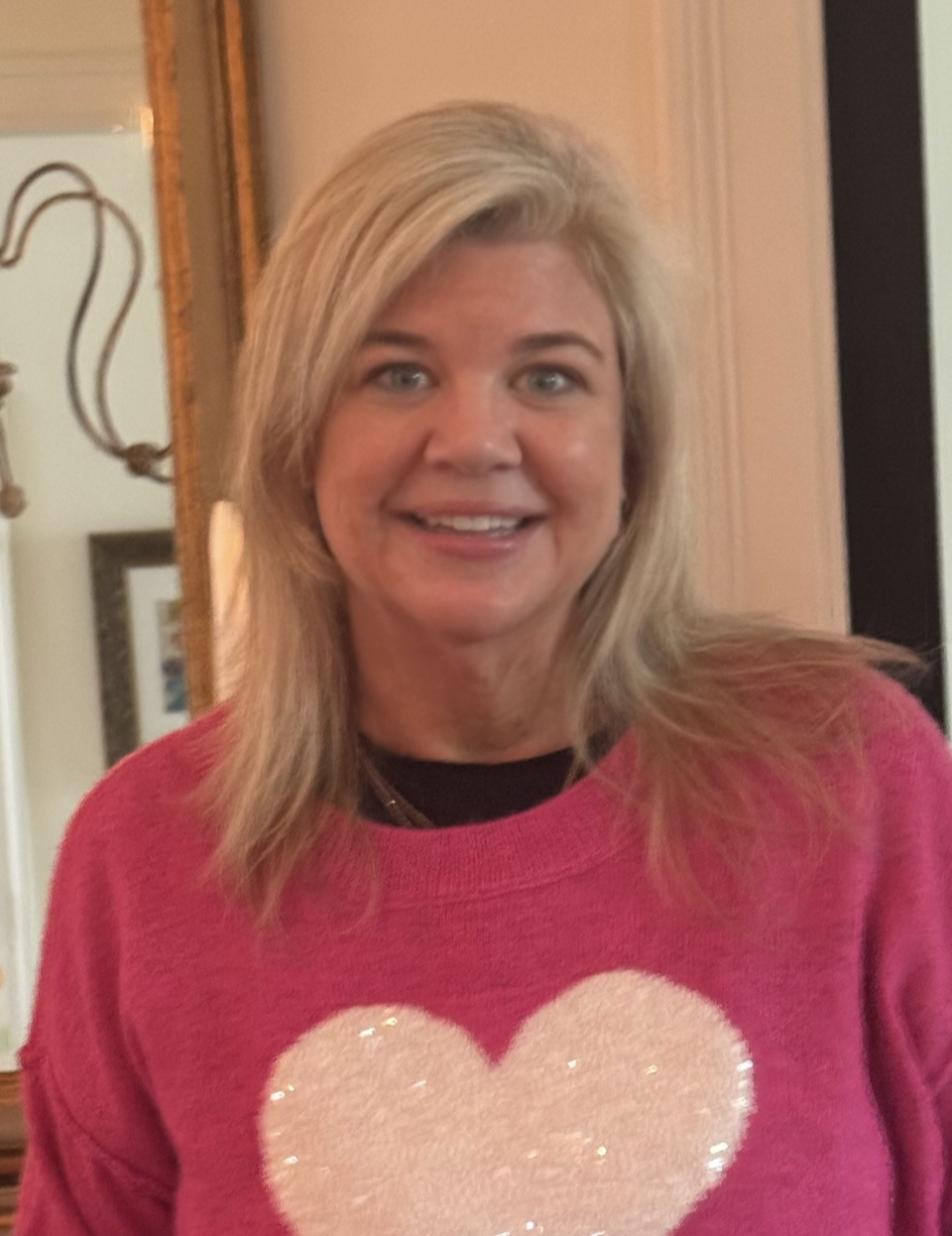 A woman with blonde hair wearing a pink sweater with a sequin heart on it, smiling indoors.
