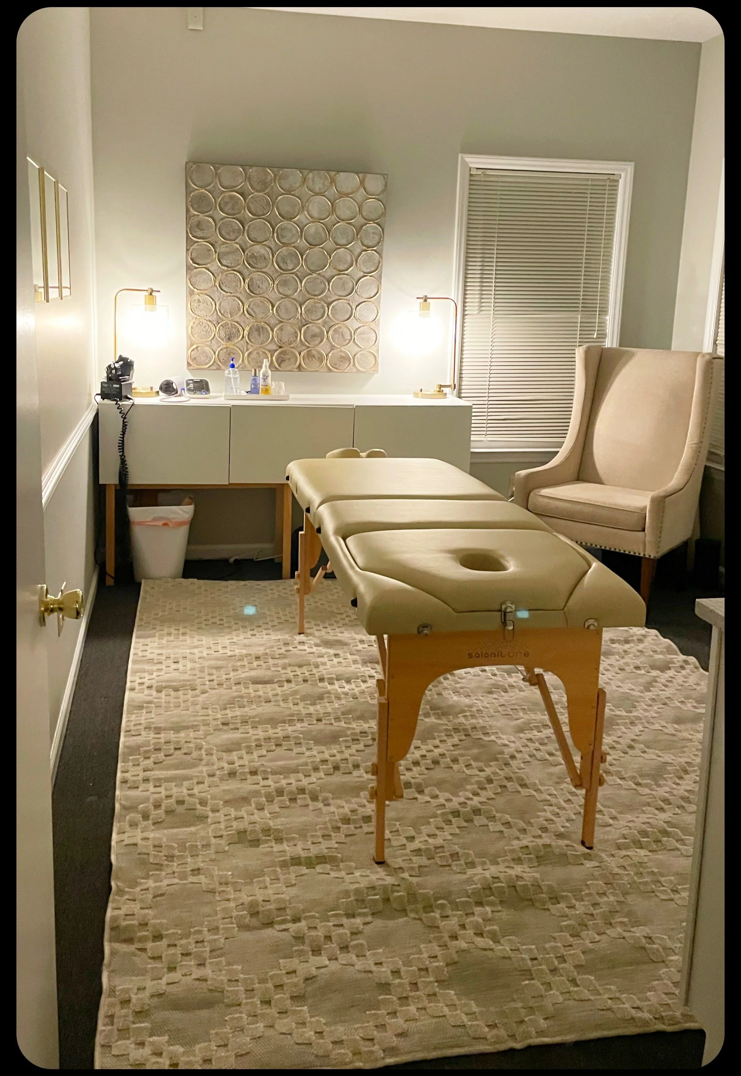 Room with a chiropractic table, beige armchair, wall art, and a side cabinet with  supplies.