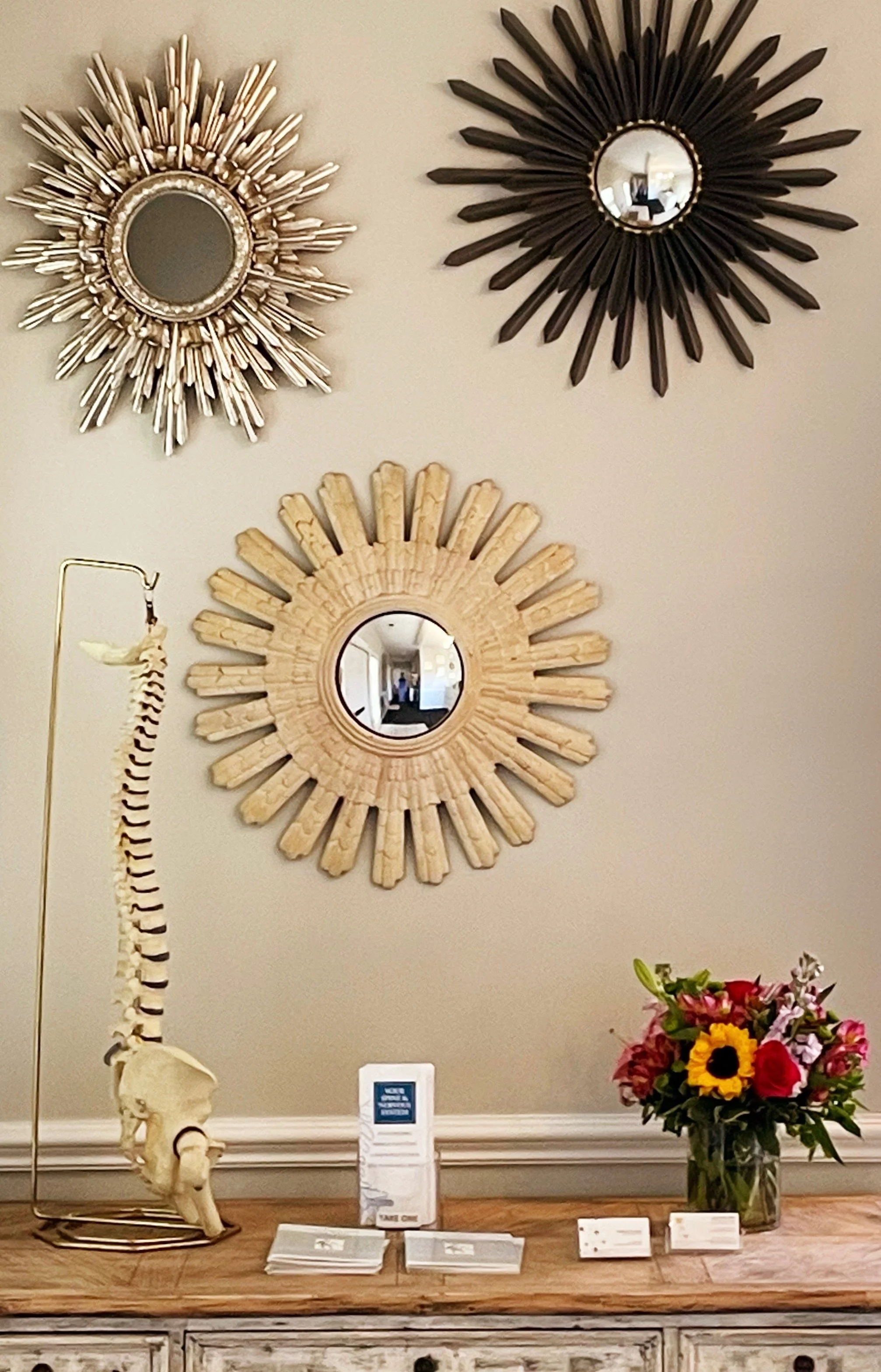 A display with three decorative mirrors and a skeleton model of a human spine on a wooden table. There is a vase with colorful flowers and some informational pamphlets on the table.