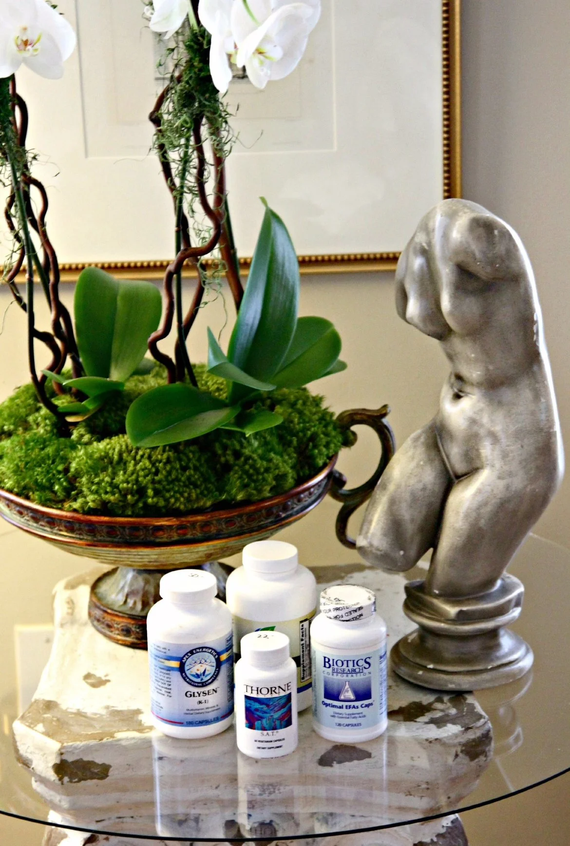 A decorative table arrangement includes a floral display of white orchids in a pedestal bowl, a stone sculpture of a torso, and several bottles of supplements on a marble surface.
