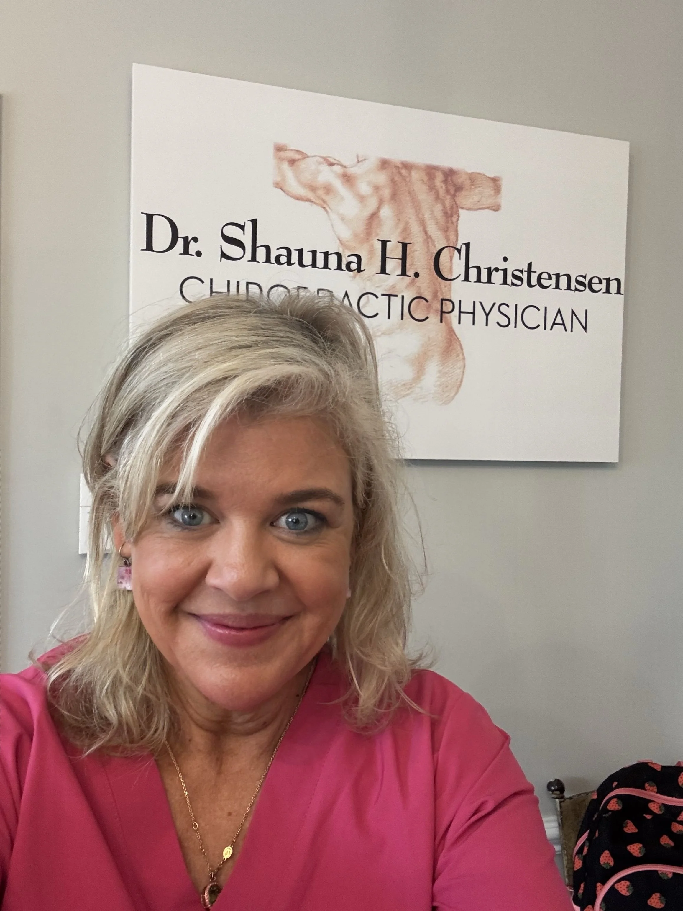 A woman with blonde hair and blue eyes smiling, wearing a pink shirt and gold jewelry in front of a sign that reads 'Dr. Shauna H. Christensen Chiropractic Physician' with a drawing of a spine.
