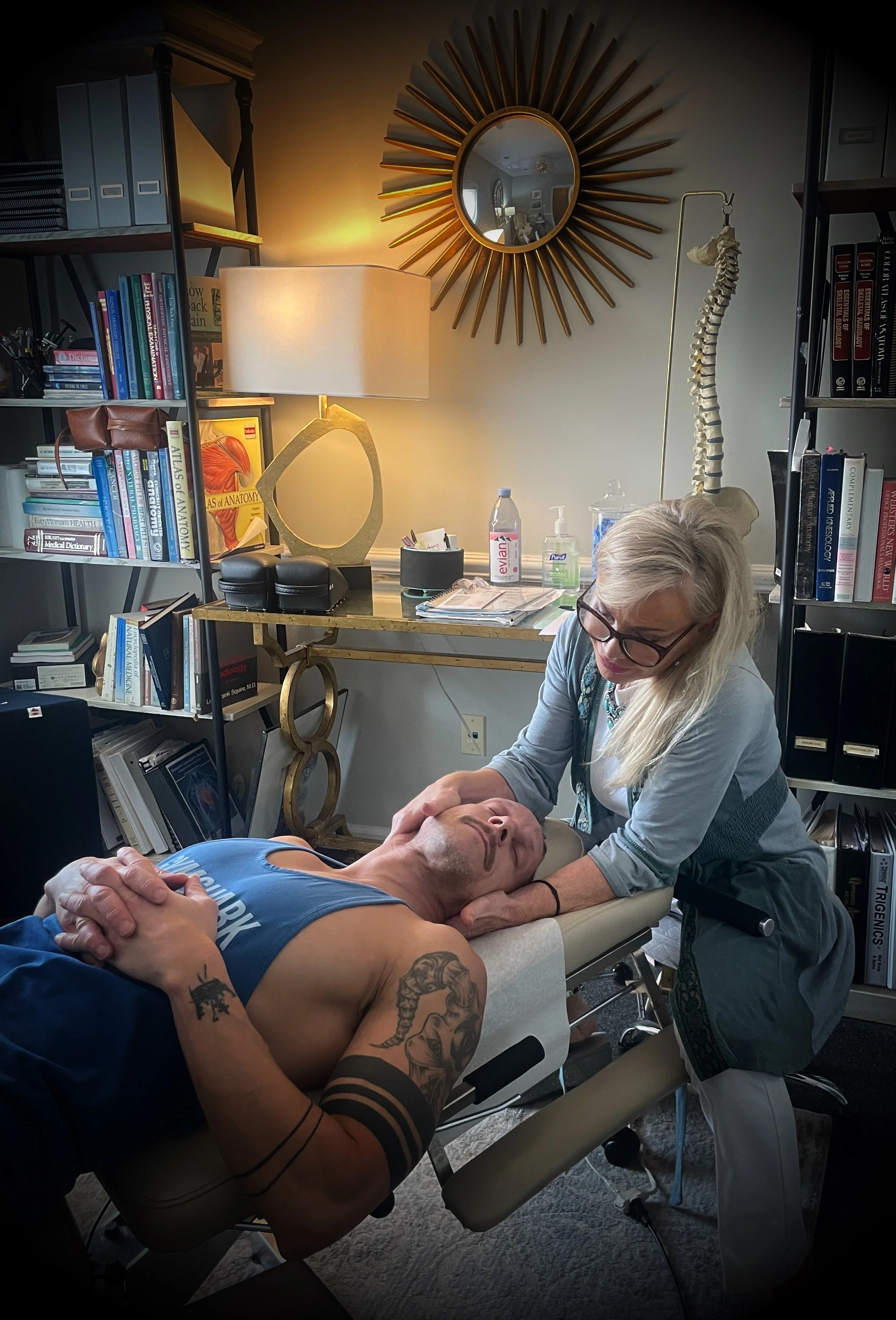 A healthcare professional performs treatment on a man lying on a treatment table in a room filled with books, medical supplies, and a large mirror.
