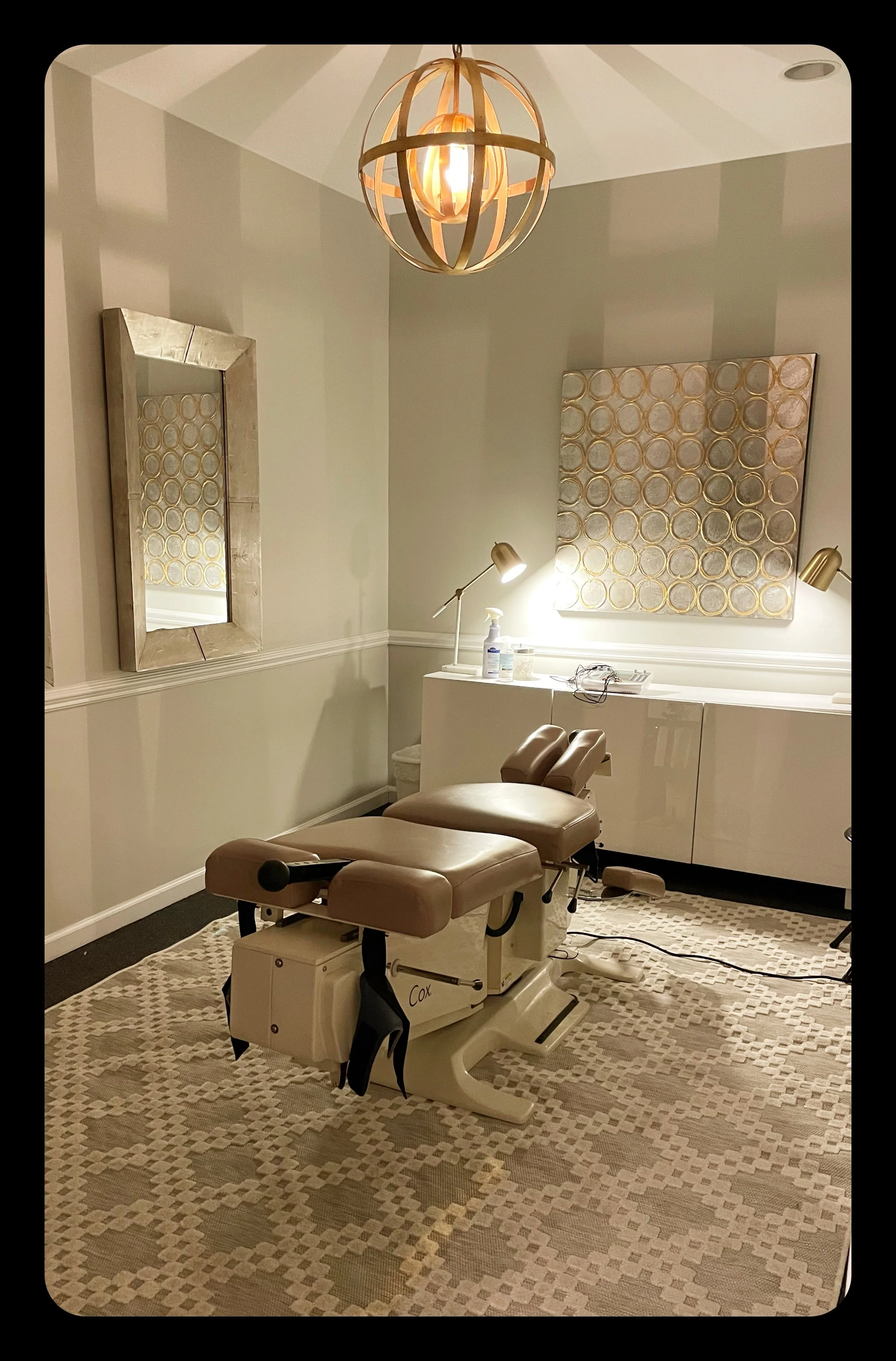 Medical examination room with an examination chair, a mirror on the wall, and a decorative circular wall art. Bright lighting and a patterned beige carpet.