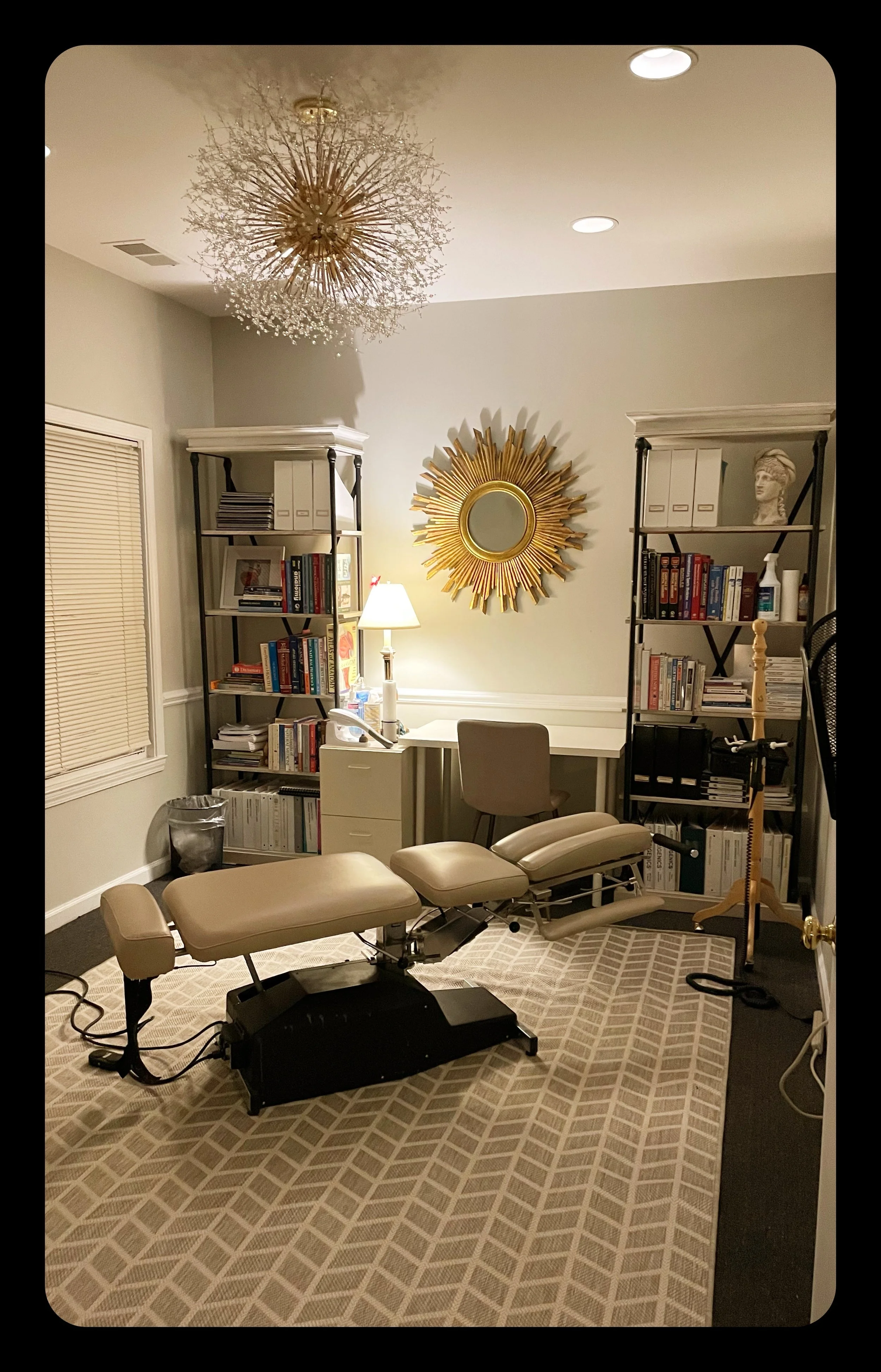 A consultation room with a chiropractic  table, bookshelves, a lamp, decorative wall mirror, and various office items.