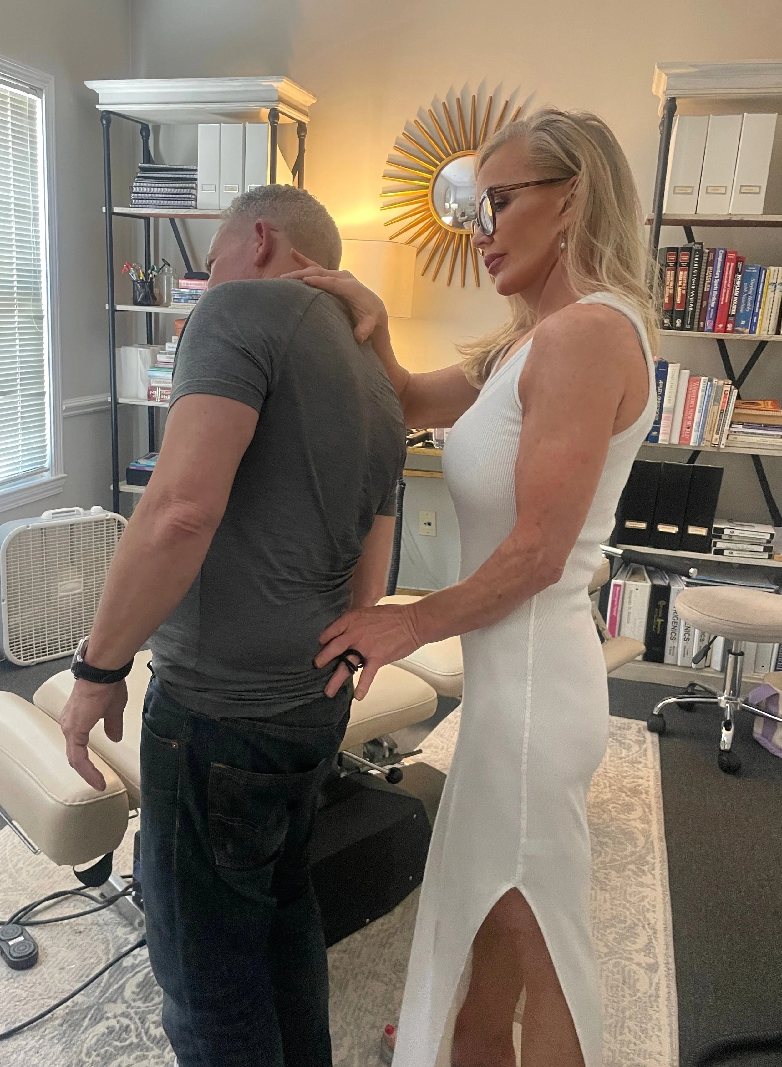 female chiropractor dressed in a white dress stands behind male patient adjusting his shoulder