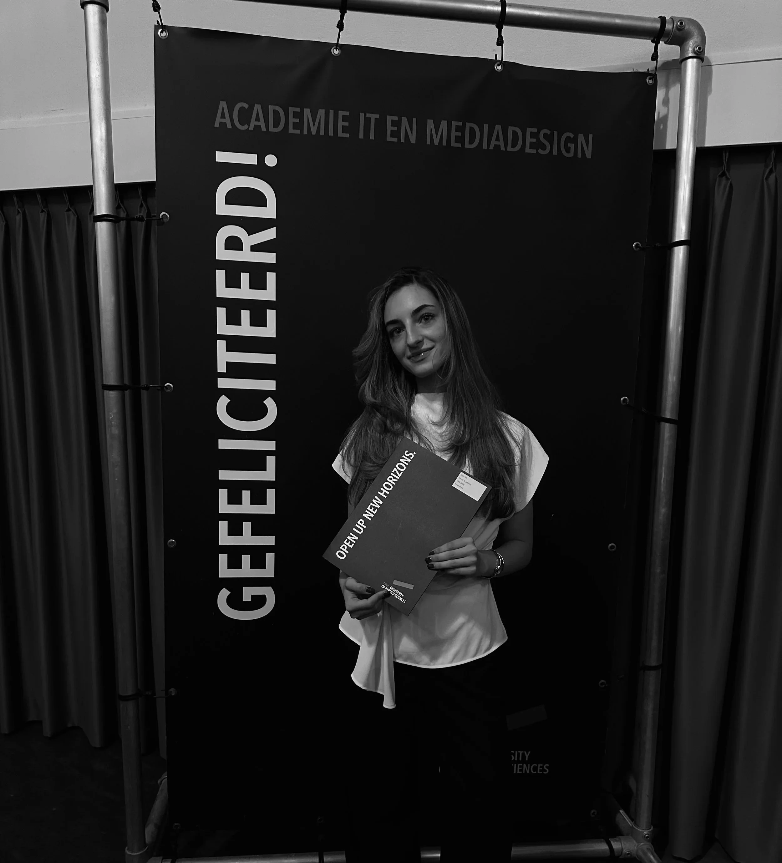 Vrouw met lang haar die een propedeuse diploma  vasthoudt met de tekst 'OPEN UP NEW HORIZONS' en poseert voor een zwart banner met de tekst 'GEFELICITEERD! ACADEMIE IT EN MEDIA DESIGN'.